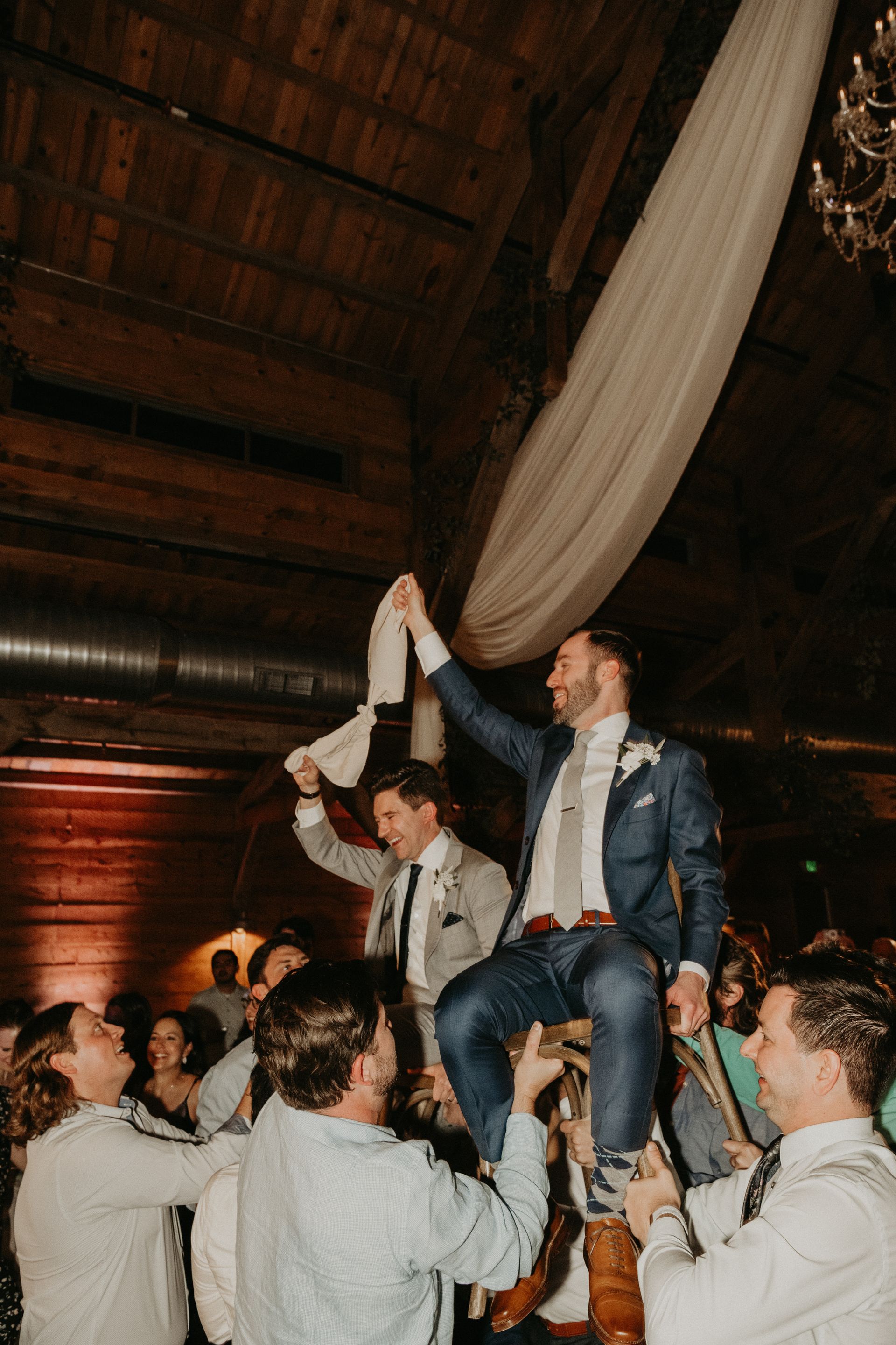 A wedding guest is carried on chairs by people in a rustic, wood-paneled venue while holding a white napkin aloft.