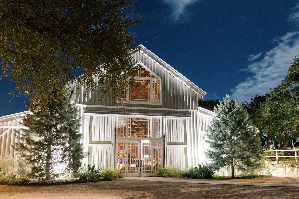 A rustic white barn illuminated at night, surrounded by trees under a dark blue sky.