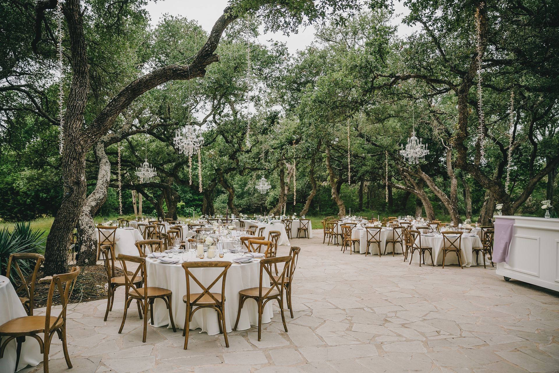 An outdoor reception area with round dining tables, wooden chairs, and crystal chandeliers hanging from oak trees.