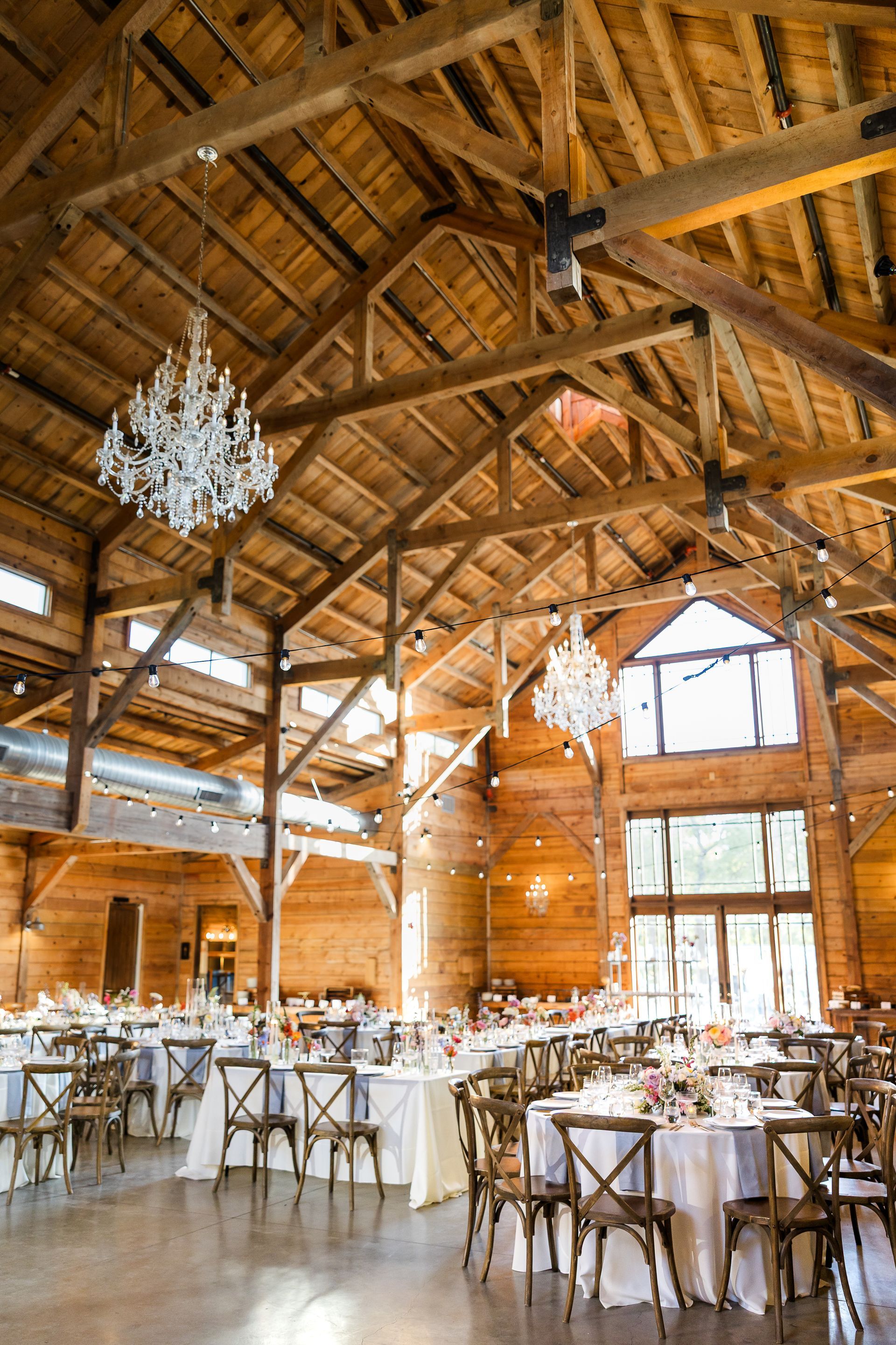 Elegant indoor wedding venue in a rustic wooden barn with exposed beams, crystal chandeliers, and set dining tables.