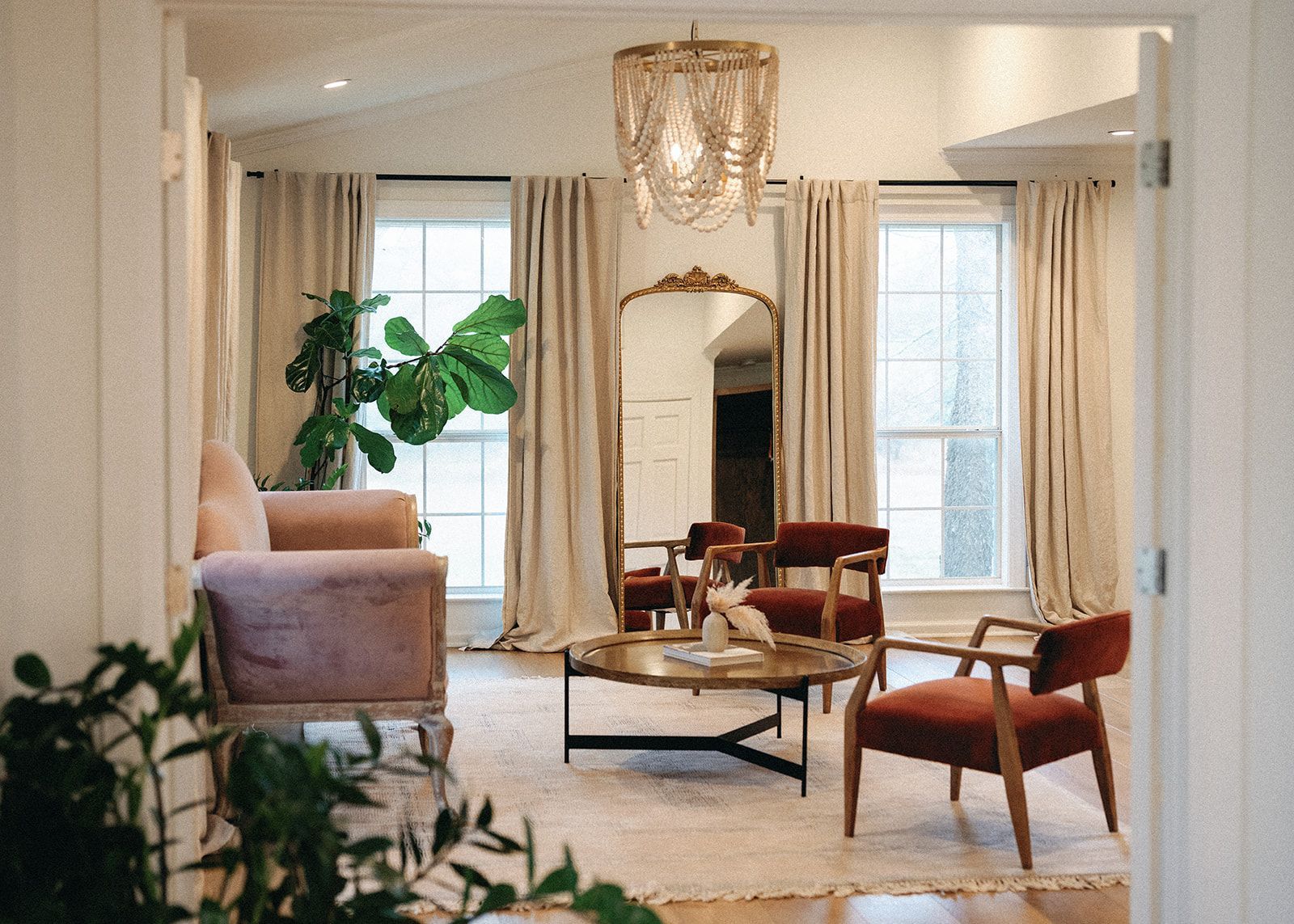 A sunlit, neutral-toned living room featuring a pink sofa, two rust-colored armchairs, a gold coffee table, and a mirror.