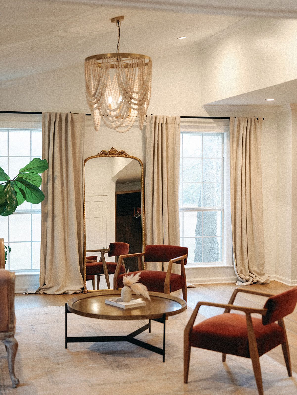 A living room featuring a round wooden coffee table, two rust-colored armchairs, a large ornate mirror, and a bead chandelier.