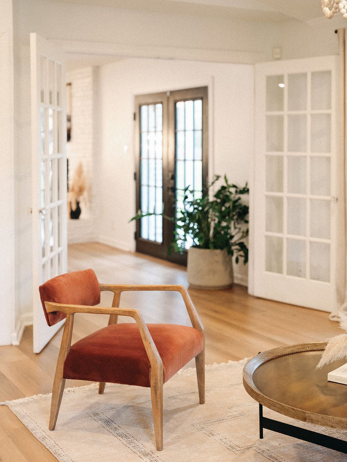 A rust-colored velvet chair sits on a textured rug in a bright, white living room with French doors and a potted plant.