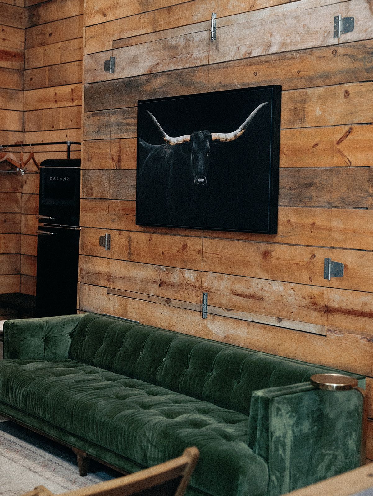 A plush dark green tufted sofa sits against a rustic wood-plank wall featuring a large framed portrait of a longhorn.