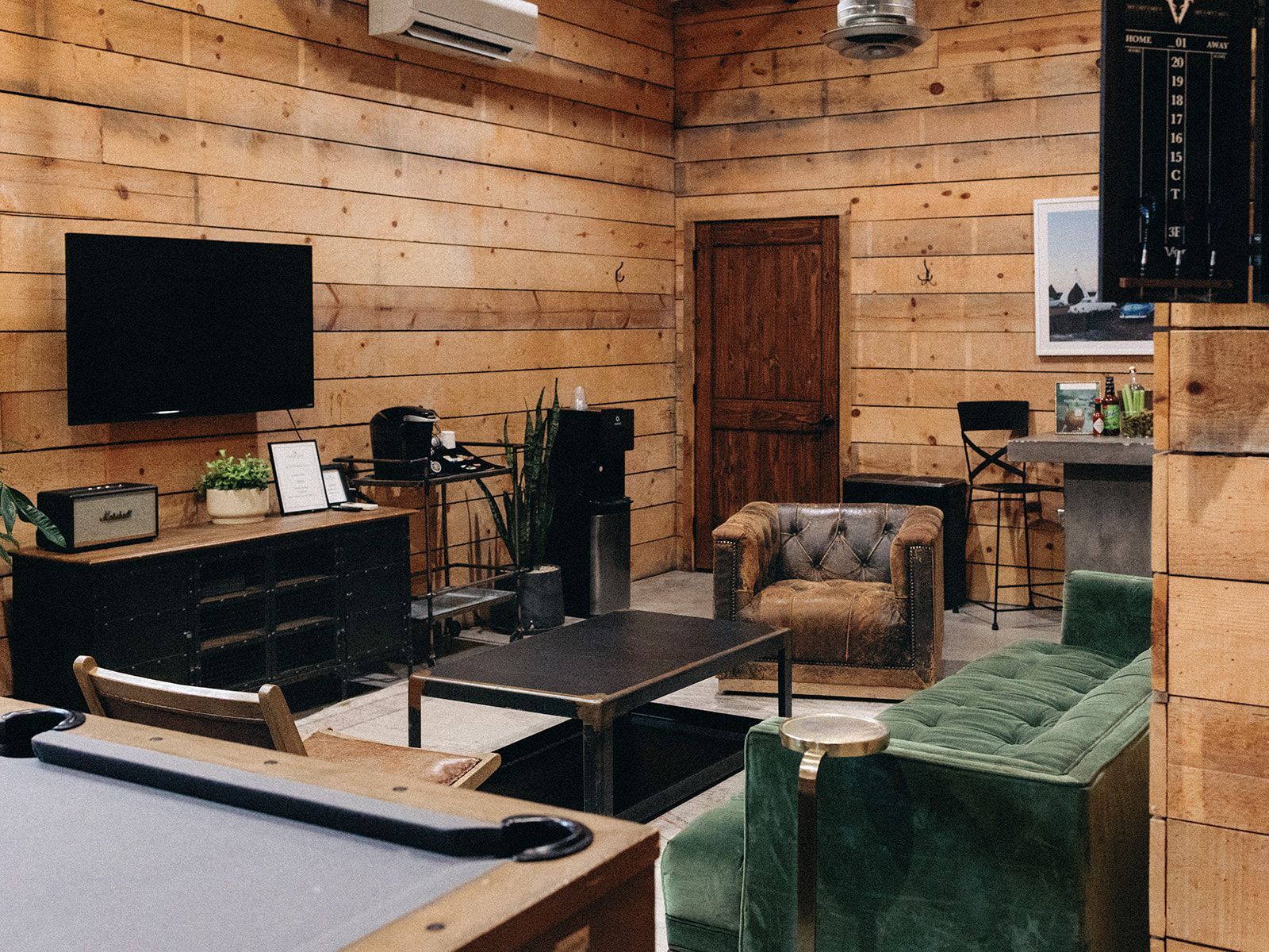A rustic lounge with log walls featuring a pool table, a green sofa, a leather armchair, a TV, and a bar area.