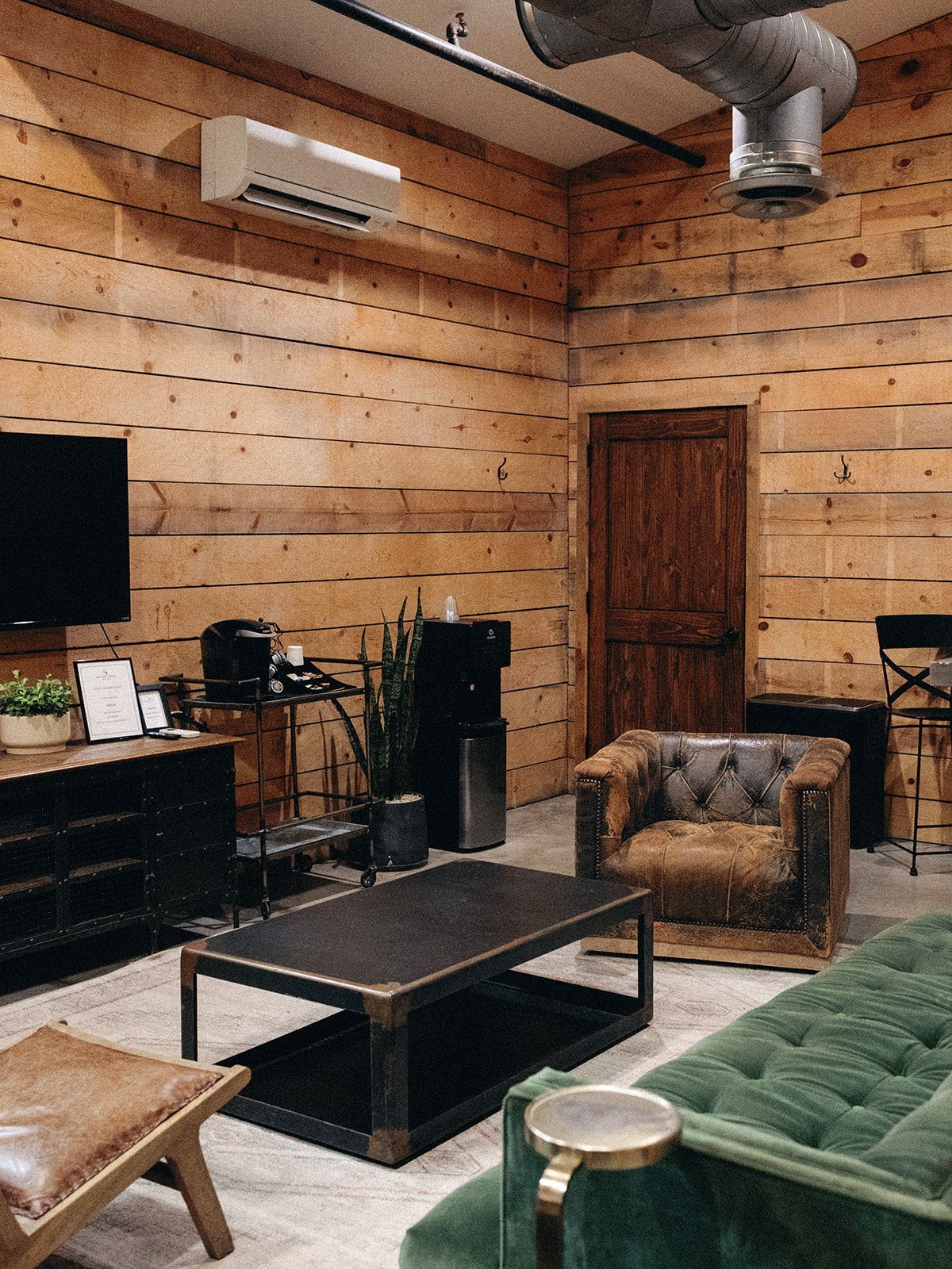 A rustic lounge with log walls, a green tufted sofa, a leather armchair, a dark coffee table, and an air conditioning unit.