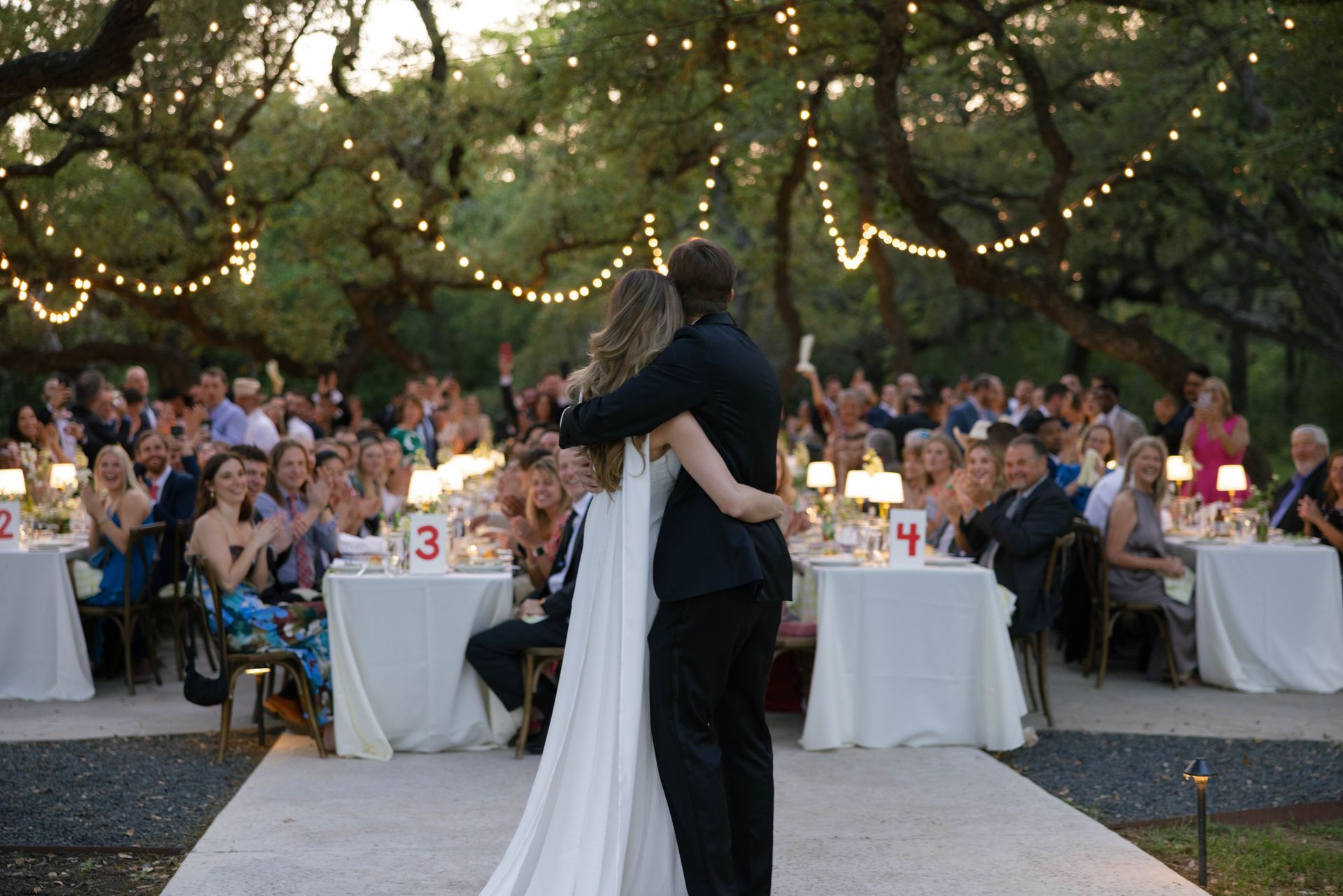 A couple dancing at an outdoor wedding reception with guests seated at tables under string lights and trees.