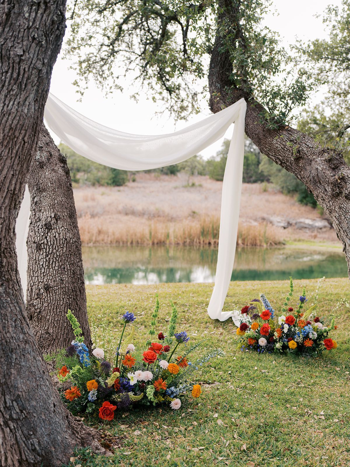 A wedding ceremony setup with white fabric draped between two trees, overlooking a pond, with two colorful floral displays.