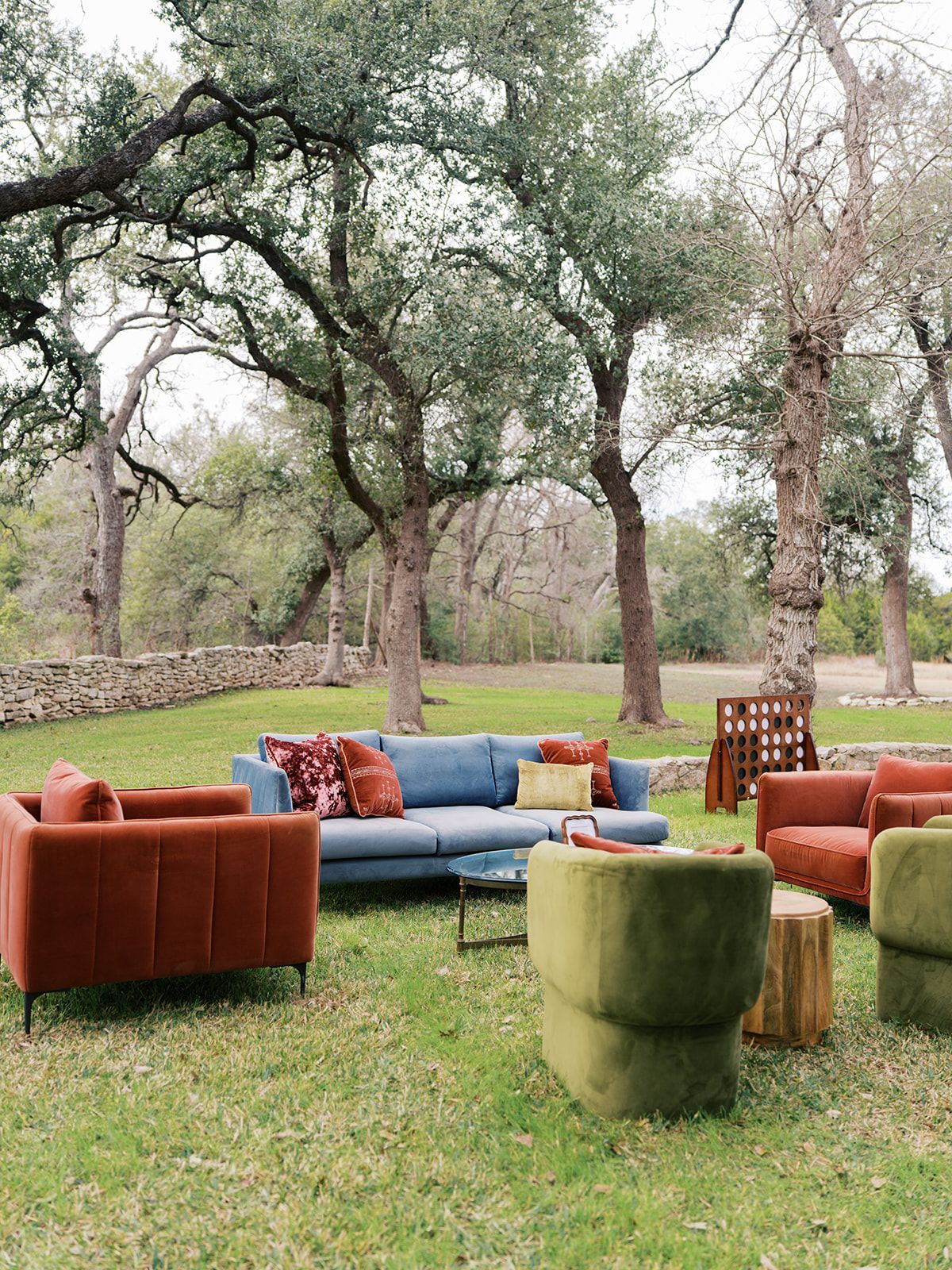 An outdoor lounge set with a blue sofa, rust-colored armchairs, and a green velvet chair on a grassy lawn under trees.
