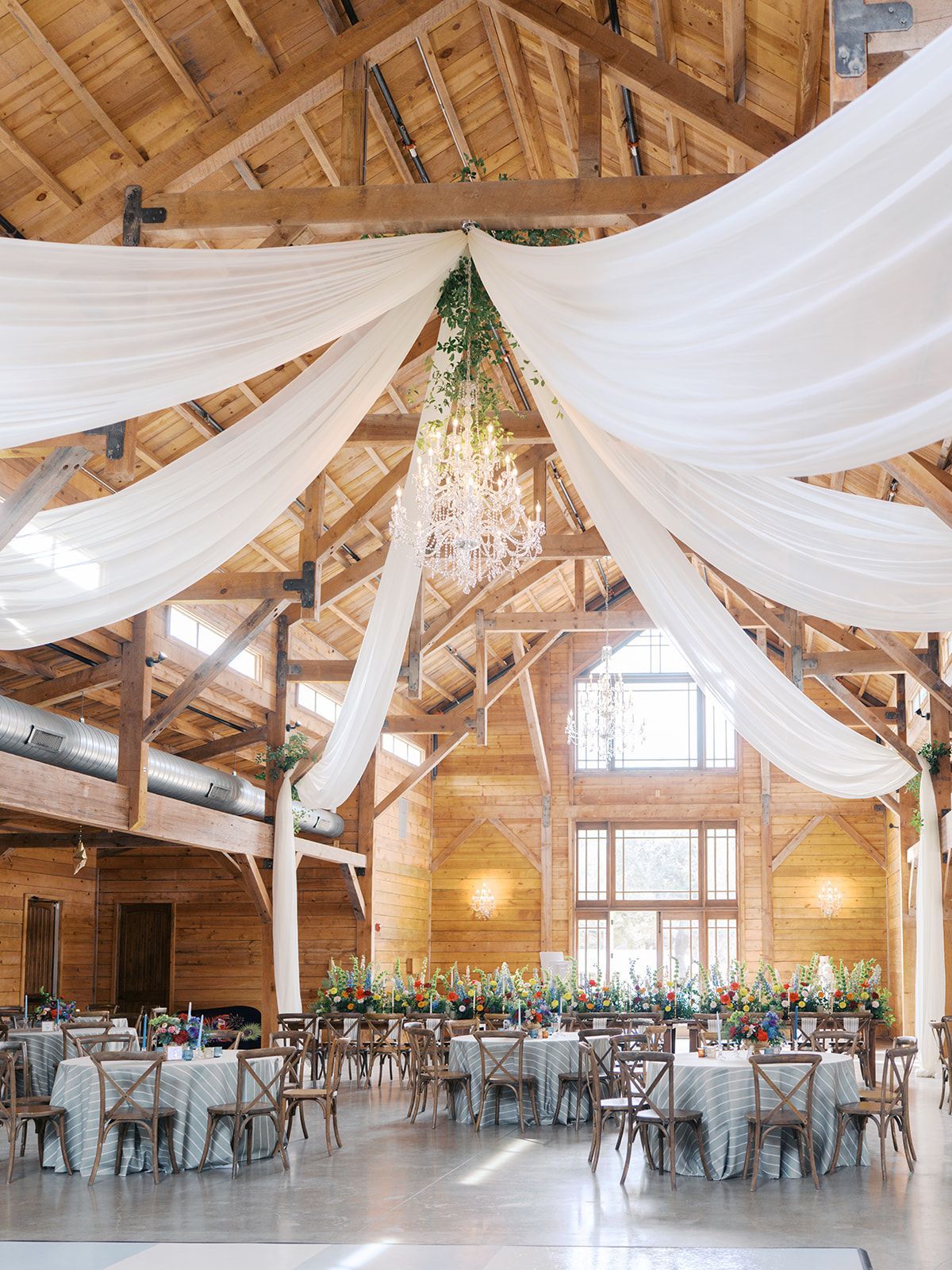 A rustic barn wedding reception with wooden beams, draped white ceiling fabric, a crystal chandelier, and tables set up.