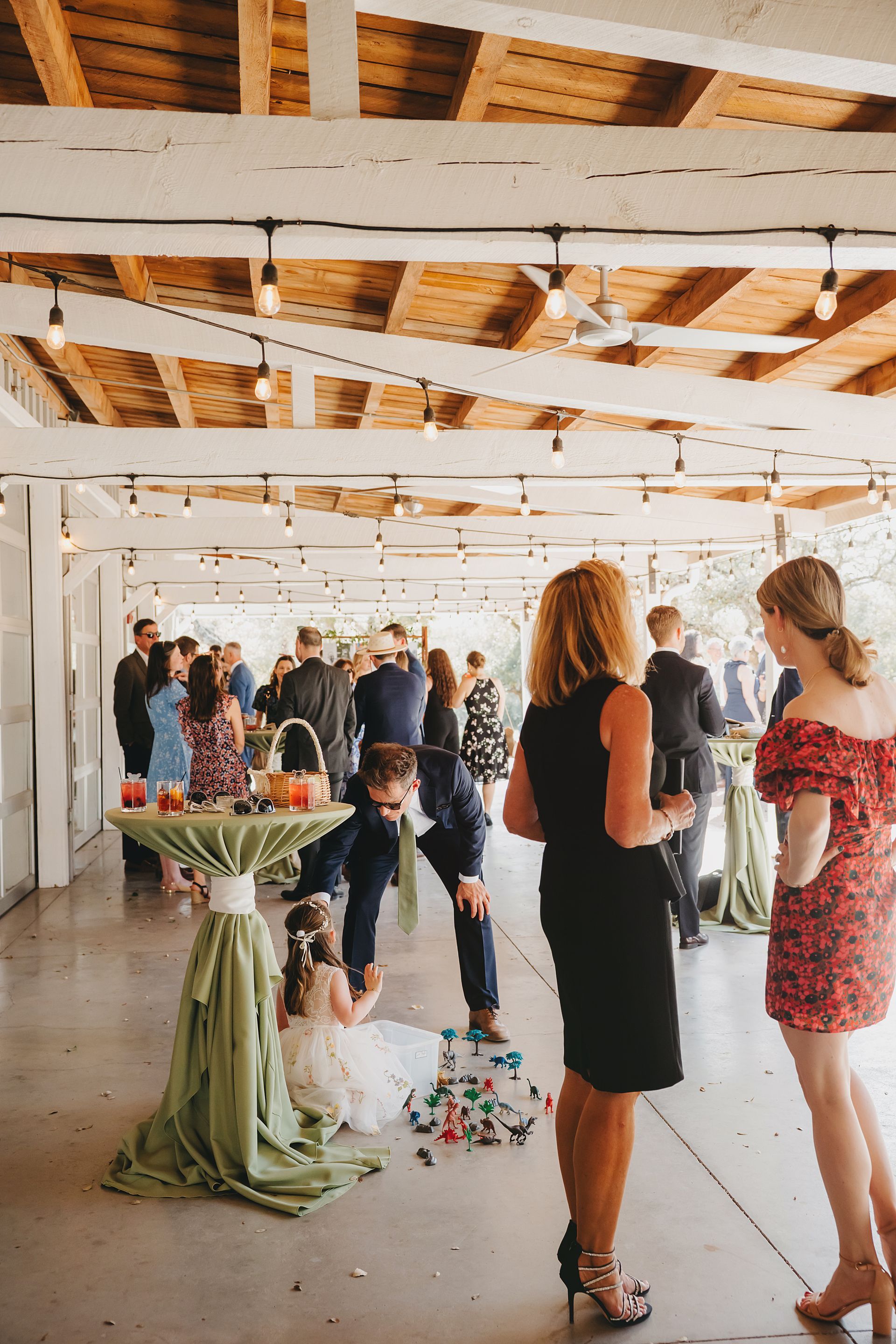 Guests socialize at an outdoor covered event space, while a person leans down to interact with a child near a table.