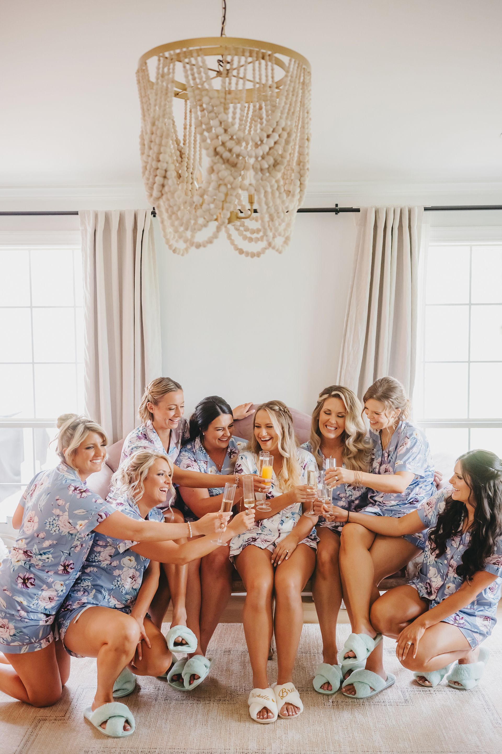 A group of friends in matching floral pajamas cheers with champagne glasses while sitting together in a bright room.