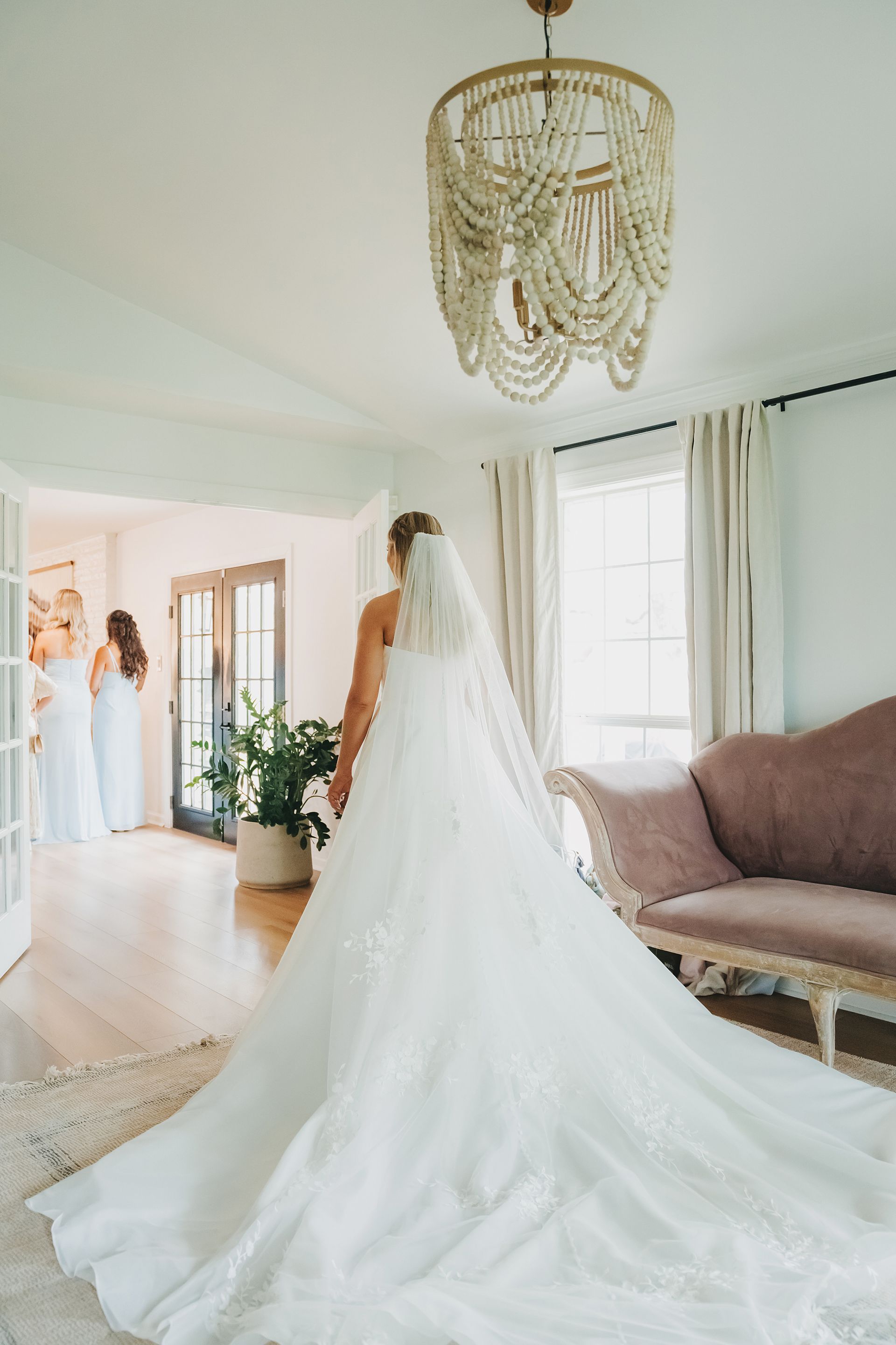 A bride in a long white gown and veil stands in a room with a chandelier, looking toward bridesmaids in another room.