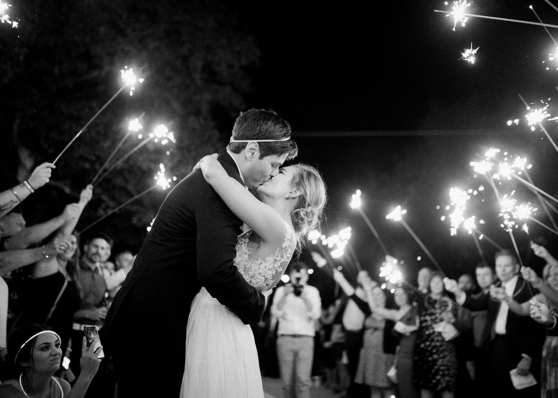 A bride and groom kiss during their wedding exit, surrounded by guests holding glowing sparklers at night.