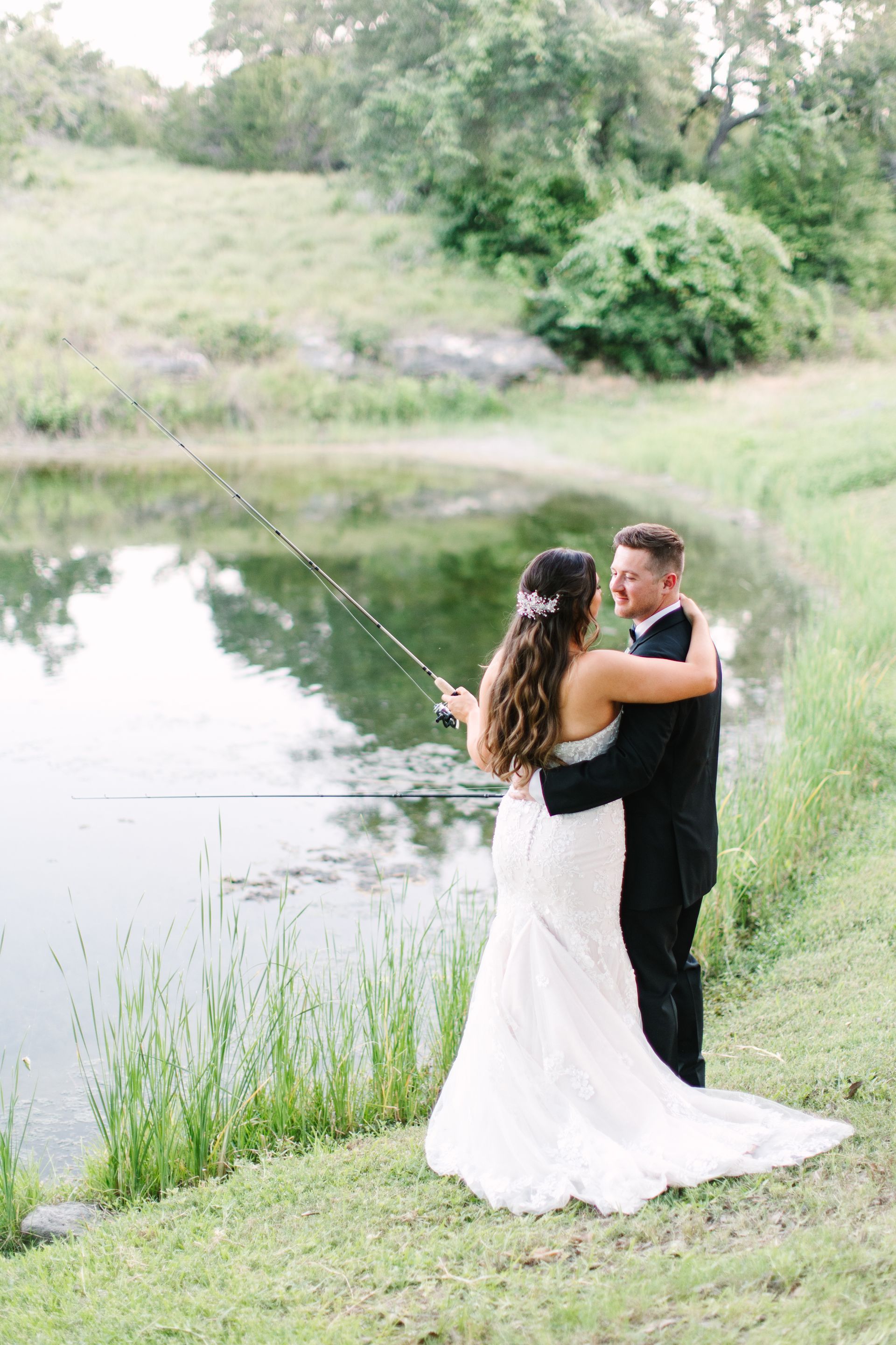 A couple in formal wedding attire stands by a pond, holding a fishing rod together while looking out over the water.