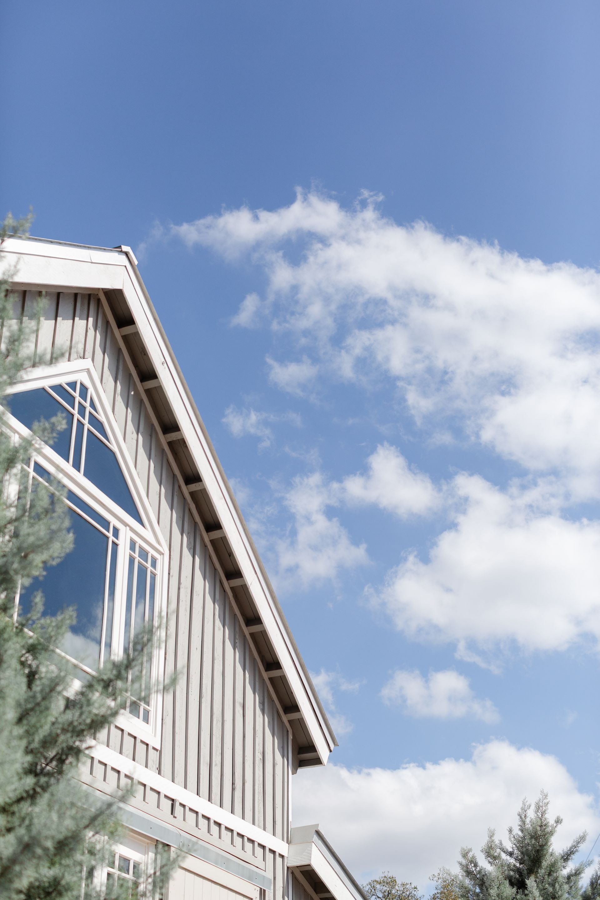 A low-angle view of a grey wooden house gable and window against a bright blue sky with soft white clouds.