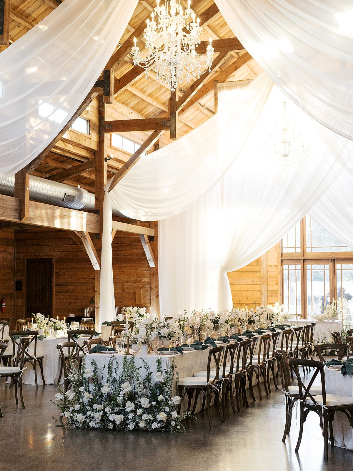 A rustic barn wedding reception with white draped fabric, a crystal chandelier, and long tables decorated with flowers.
