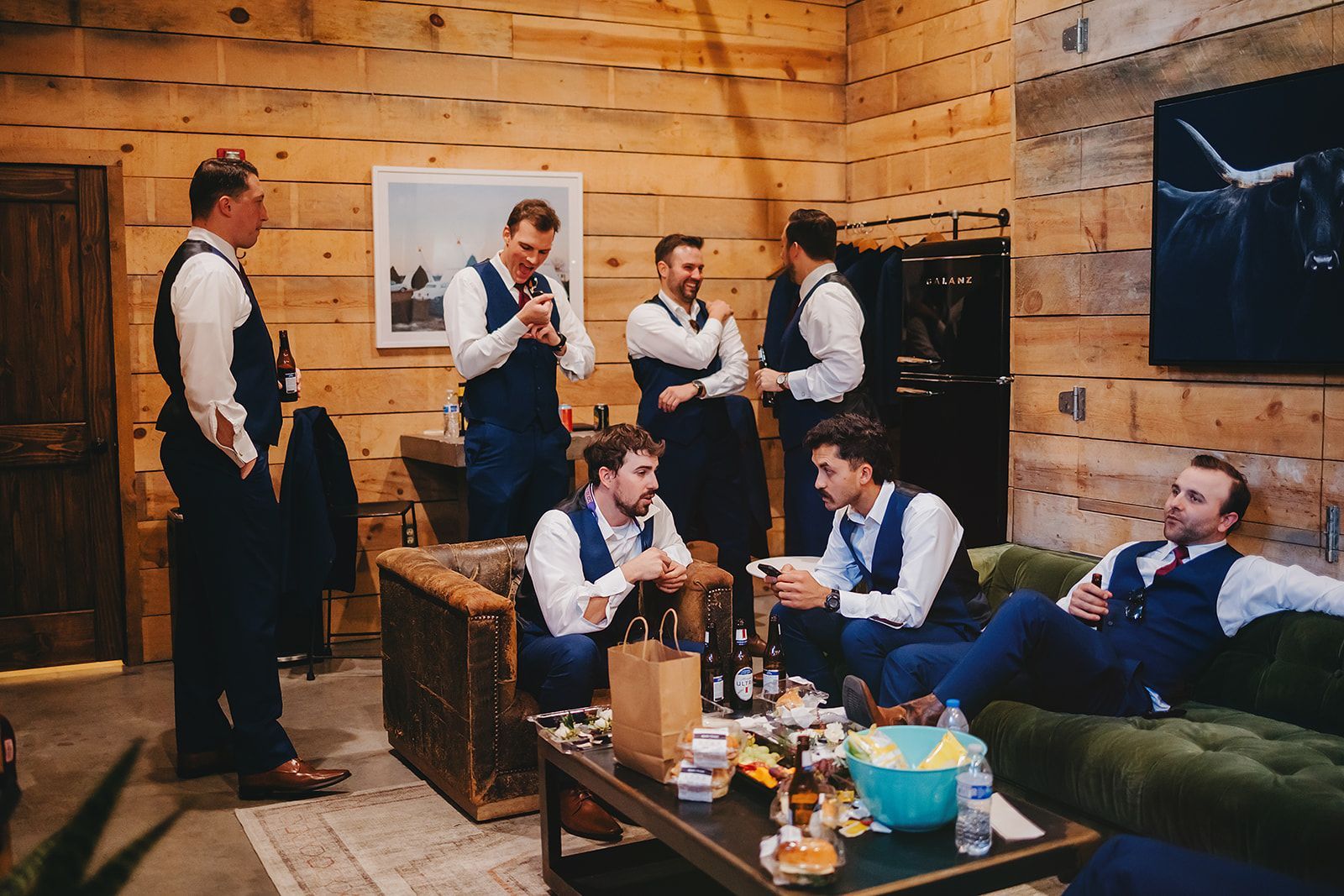 A group of men in suits and vests gather in a rustic wood-paneled room, socializing near a green velvet sofa and table.