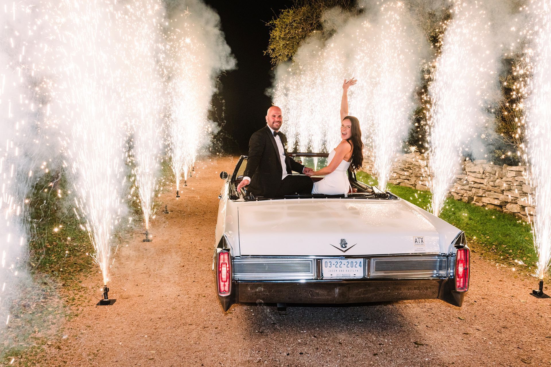 Newlyweds in a white convertible driving through a pyrotechnic display at night. Woman raises her arm; man smiles.