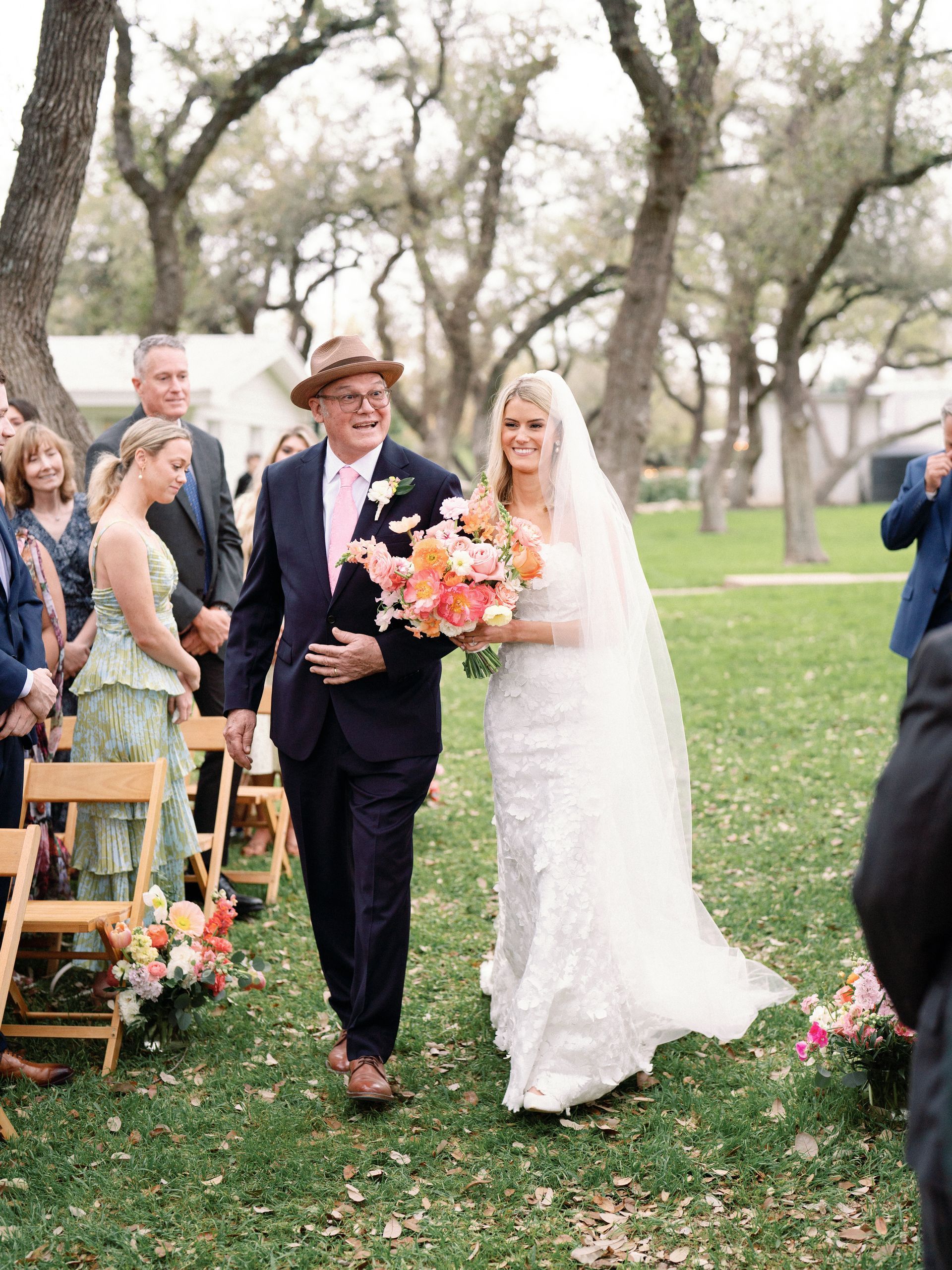 A bride in a white wedding gown and veil walks down an outdoor grassy aisle, arm-in-arm with a man in a suit and hat.