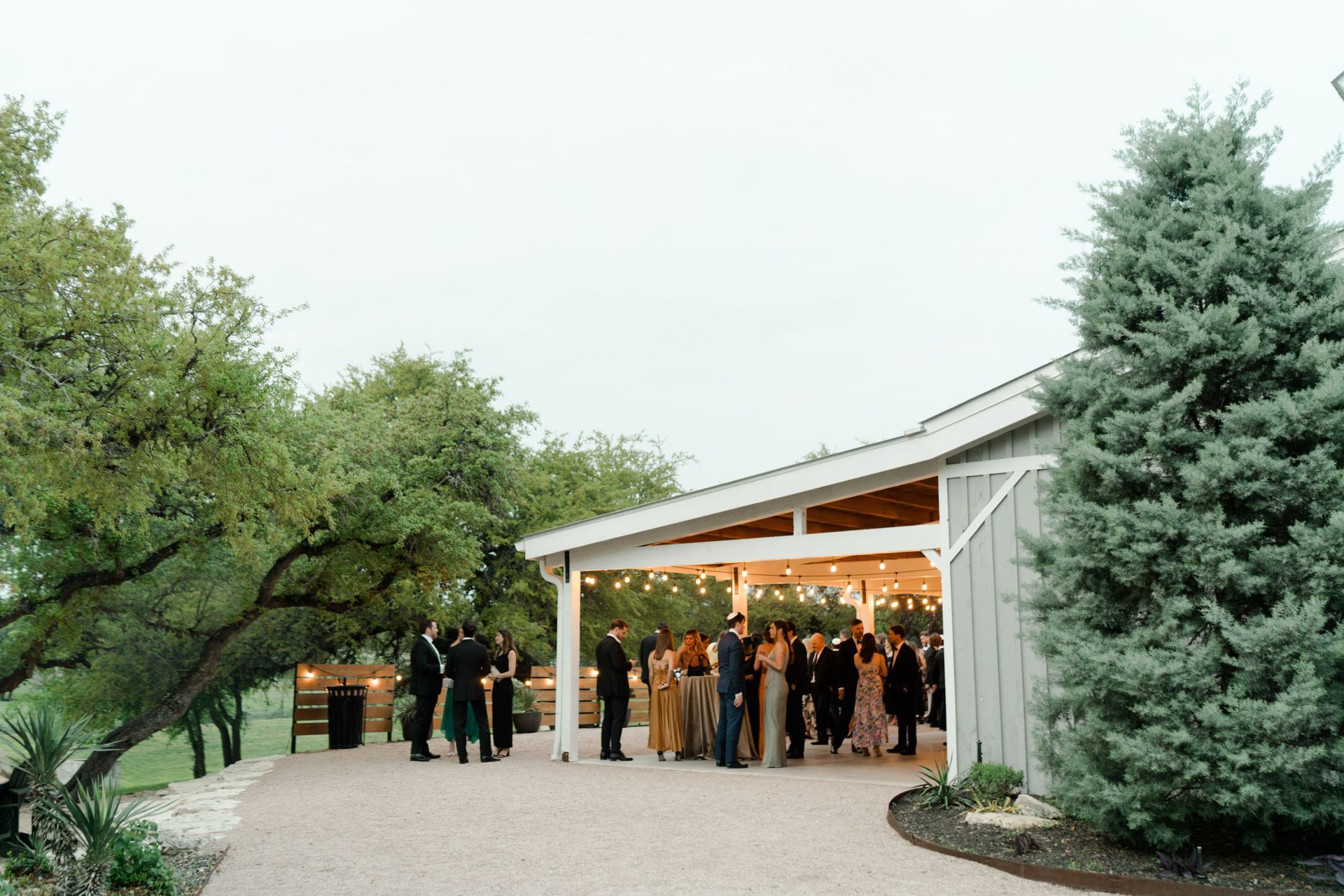 A group of people gathers for an outdoor social event under the covered patio of a rustic, grey-sided building.