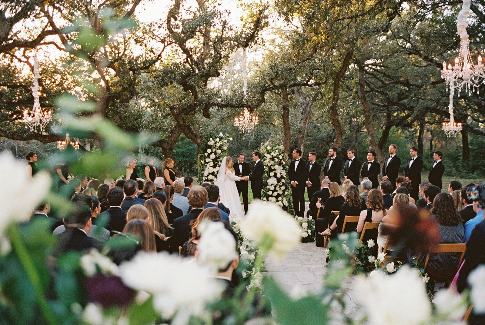 A wedding ceremony held outdoors beneath large trees with hanging chandeliers, featuring a couple at the altar.