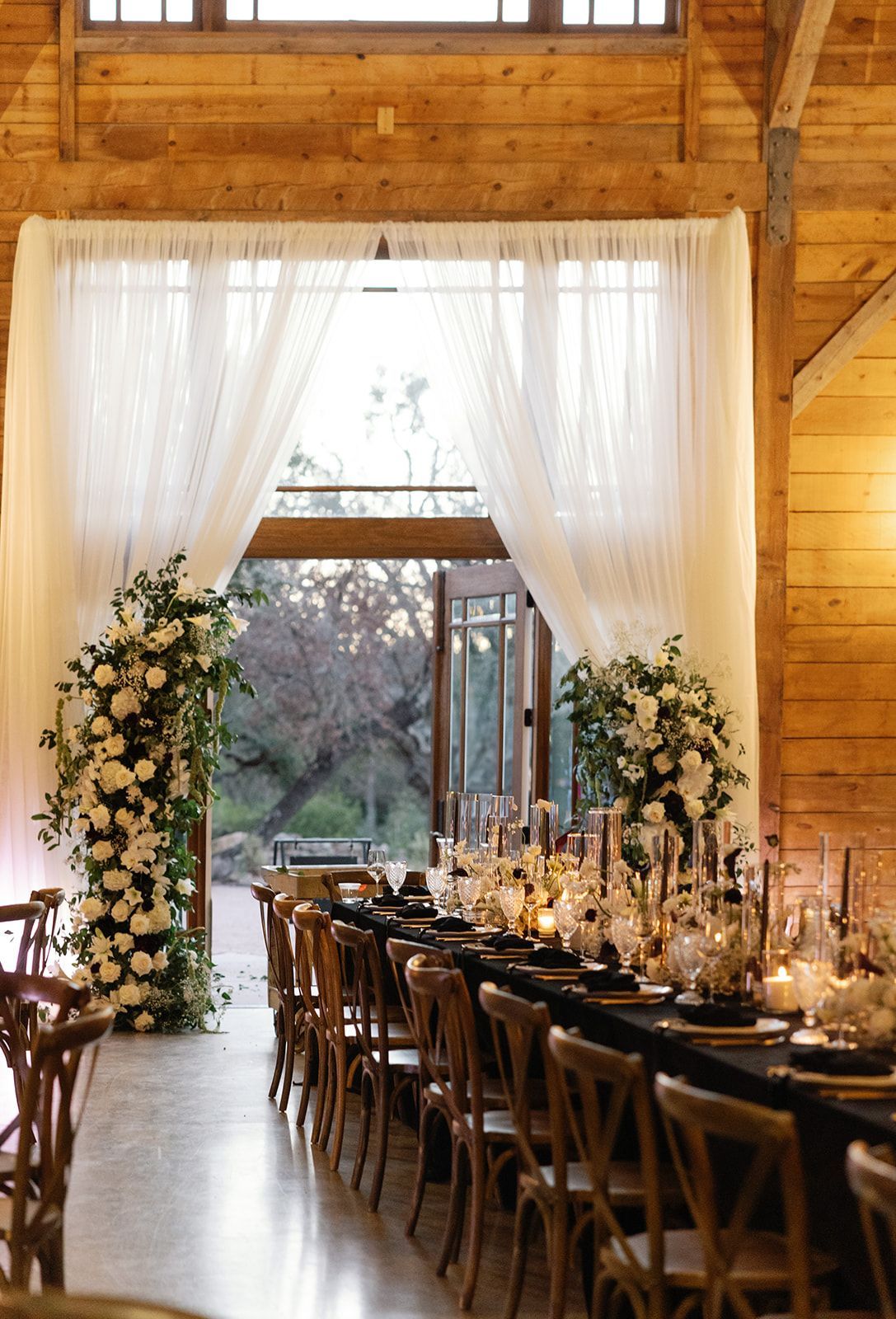 A long banquet table set for a wedding in a rustic wooden barn, featuring floral arrangements and open double doors.