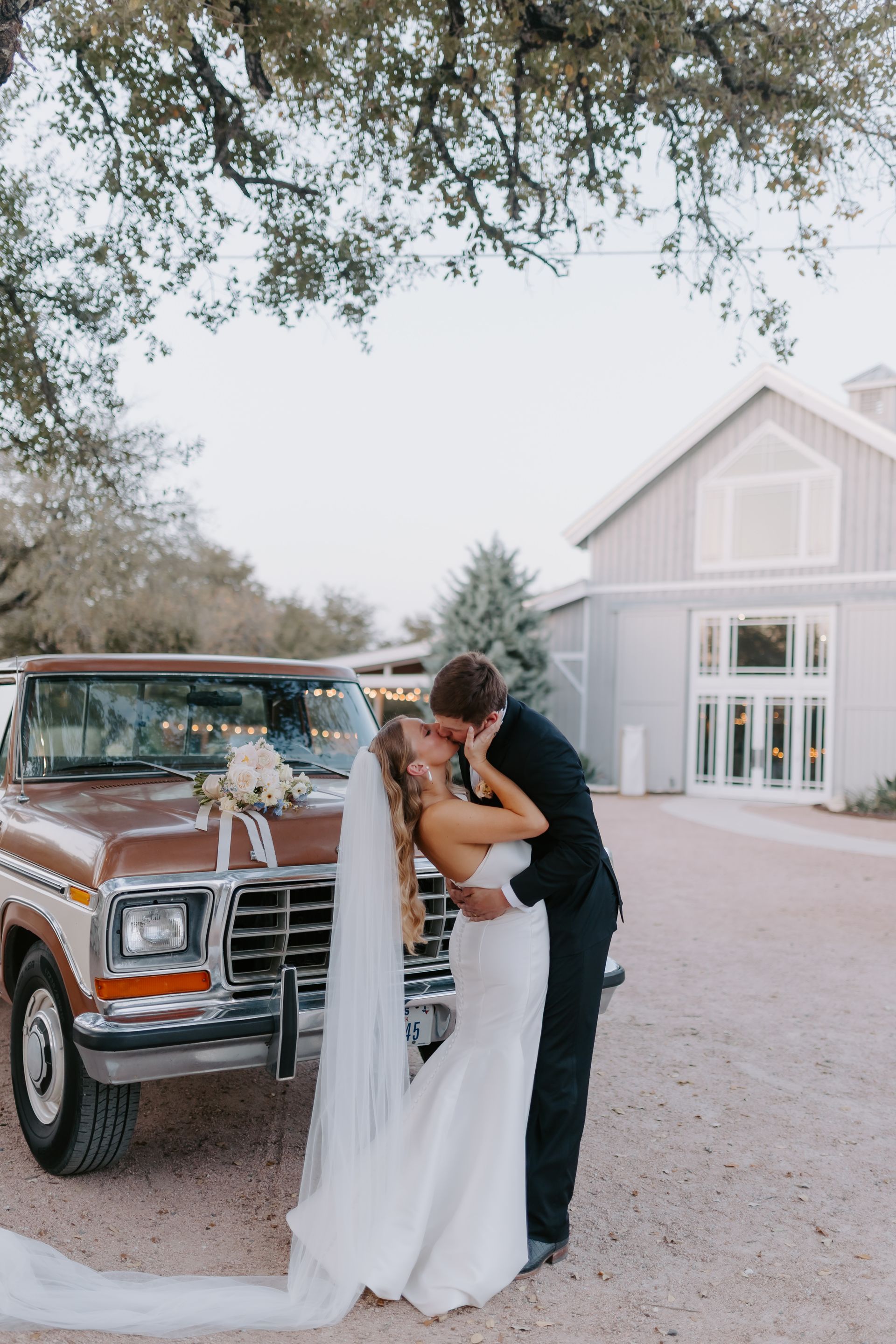 A couple in wedding attire embraces and kisses in front of a vintage brown truck at an outdoor venue.