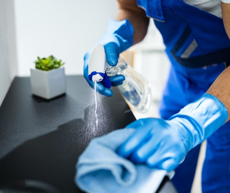 A man in blue overalls and blue gloves is cleaning a counter with a cloth and spray bottle.