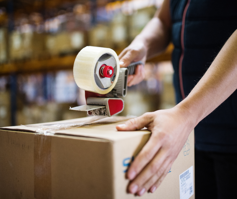 A person is taping a cardboard box in a warehouse.