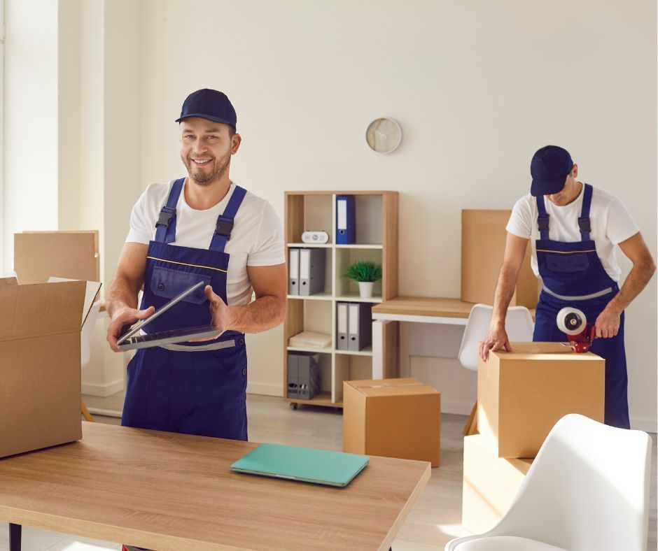 Two men in blue overalls are moving boxes in a room.