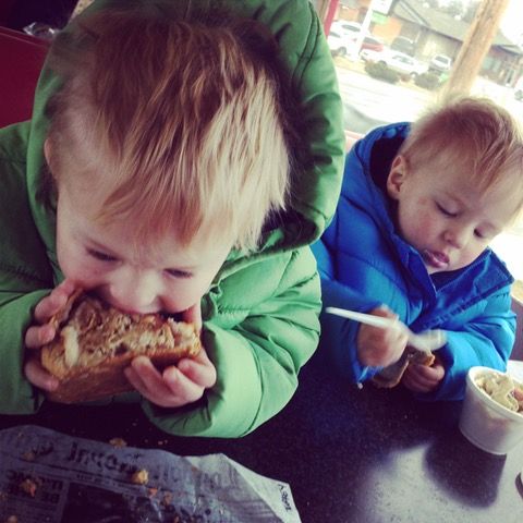 Two young boys are sitting at a table eating food and one of them is wearing a green jacket