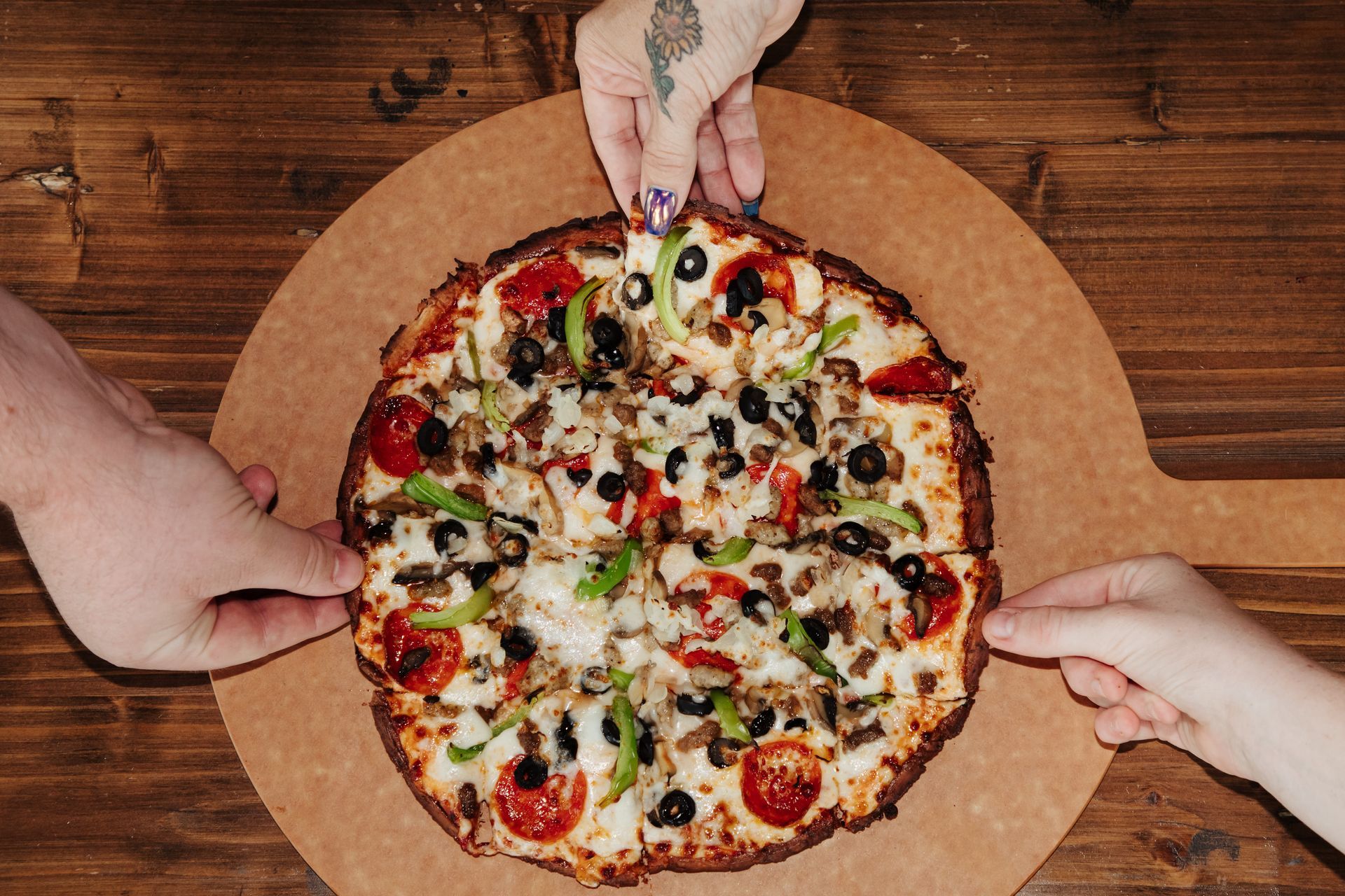 Two people are reaching for a pizza on a wooden table.