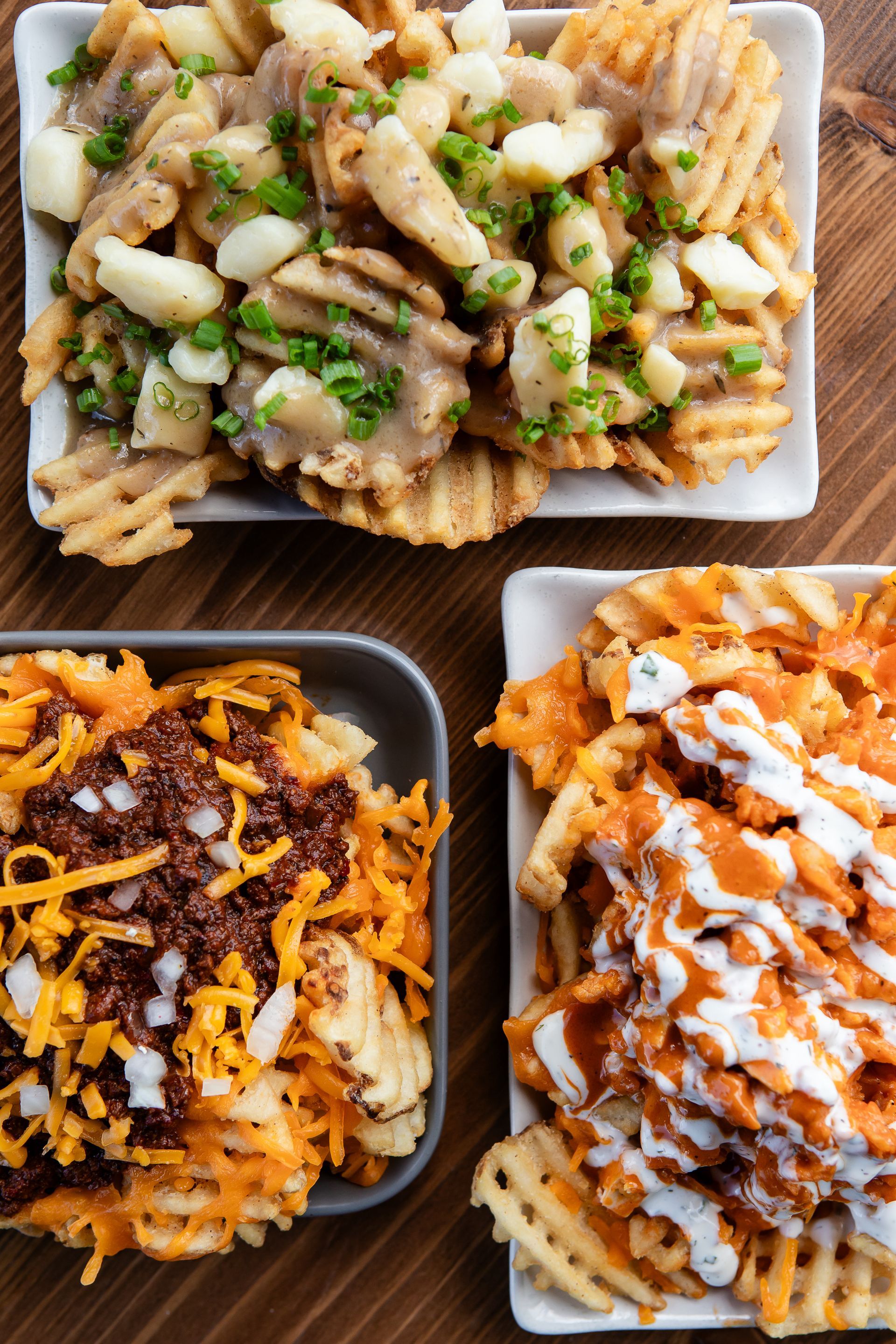 Three plates of waffle fries with different toppings on a wooden table.