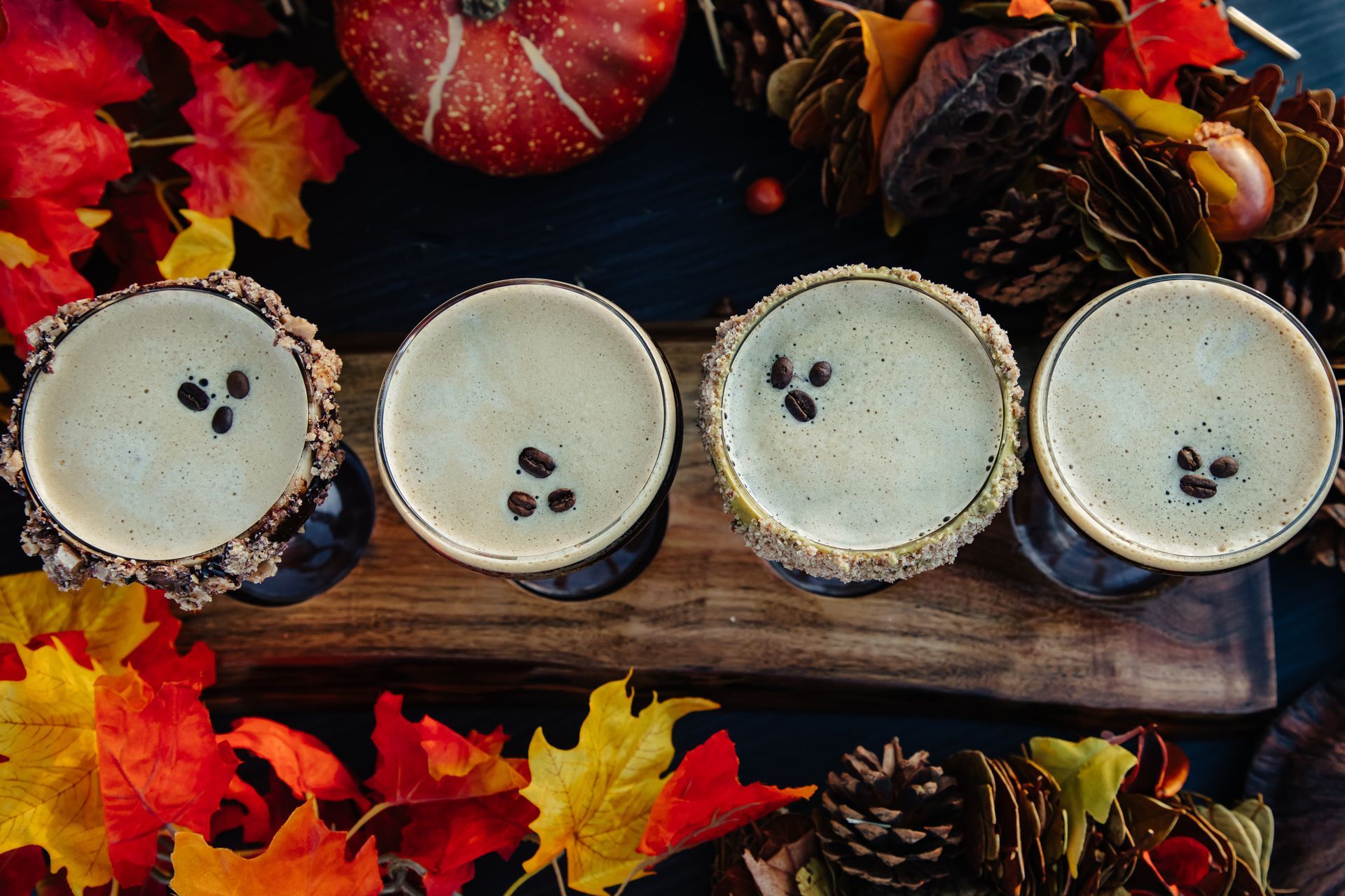Four glasses of coffee are sitting on a wooden table surrounded by leaves and pine cones.