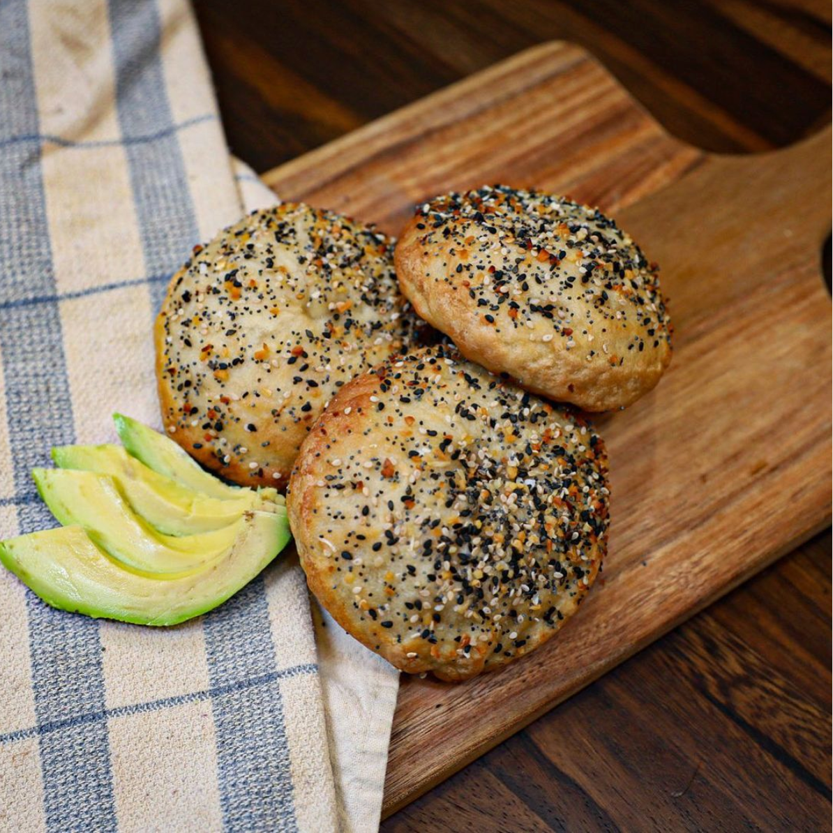 Three bagels are on a wooden cutting board next to a slice of avocado.