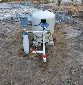 Different Kinds Of Water Pumps — Palatka, Fl — Moore’s Well Drilling Well water system with white tank, pipes, and control box outdoors on dirt.