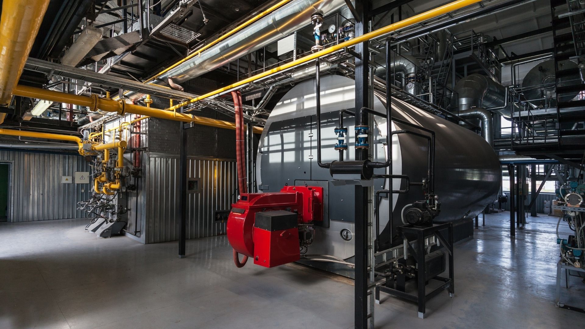 Industrial boiler room with a large black boiler, red burners, and yellow pipes.