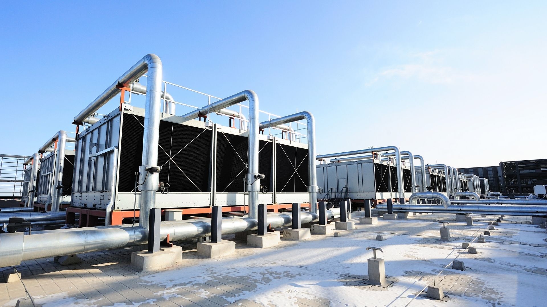 Cooling towers on a rooftop with metallic pipes and a blue sky, some snow on the ground.