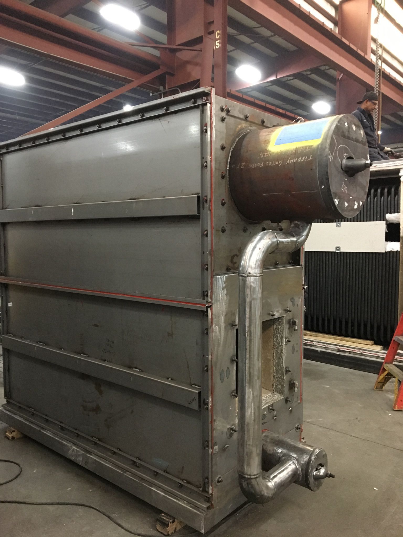 Large industrial metal box with cylindrical section and pipes attached, in a factory setting.