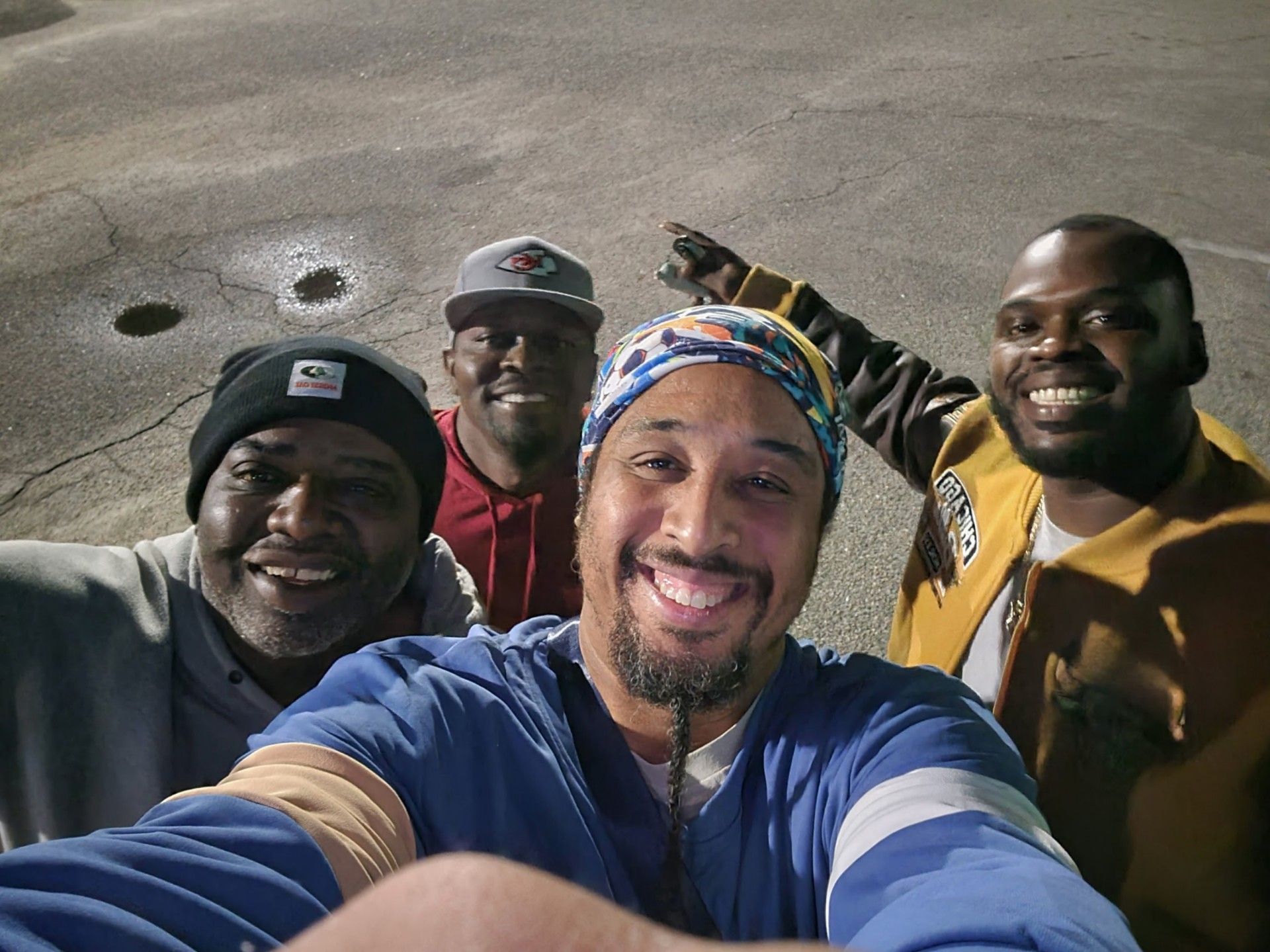Four smiling Black men take a selfie outdoors; one points up.