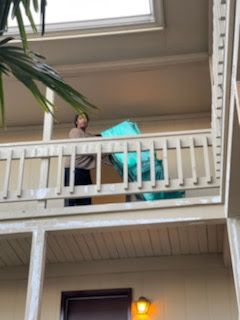Person on a balcony struggles to move a large, teal object. Beige building, overcast day.