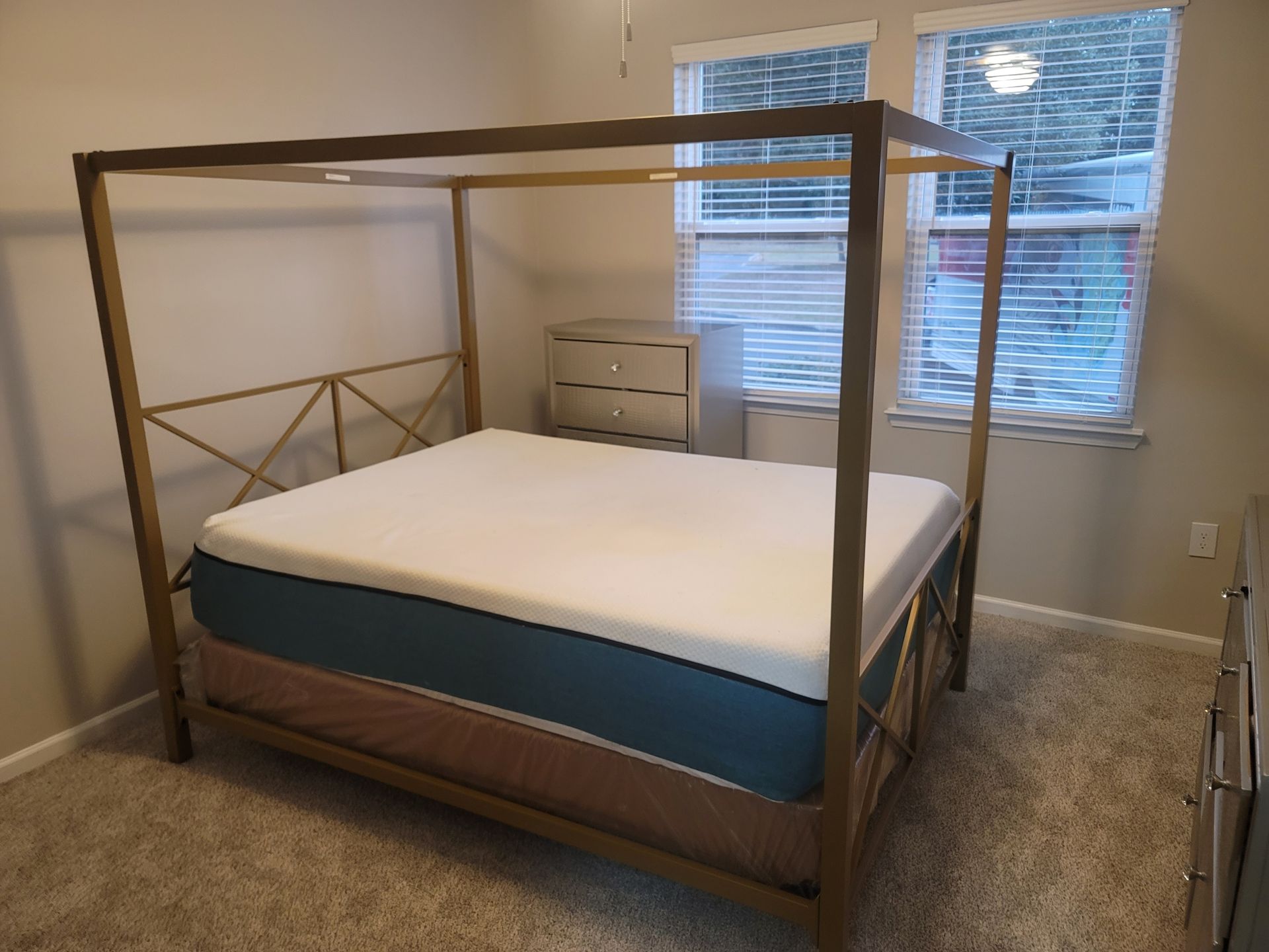 Bedroom with a gold canopy bed, a white dresser, and two windows with blinds.