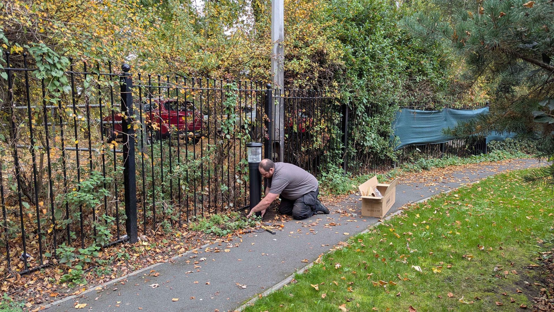 Man kneeling, raking leaves on a sidewalk next to a fence and grassy area. Cardboard box nearby.