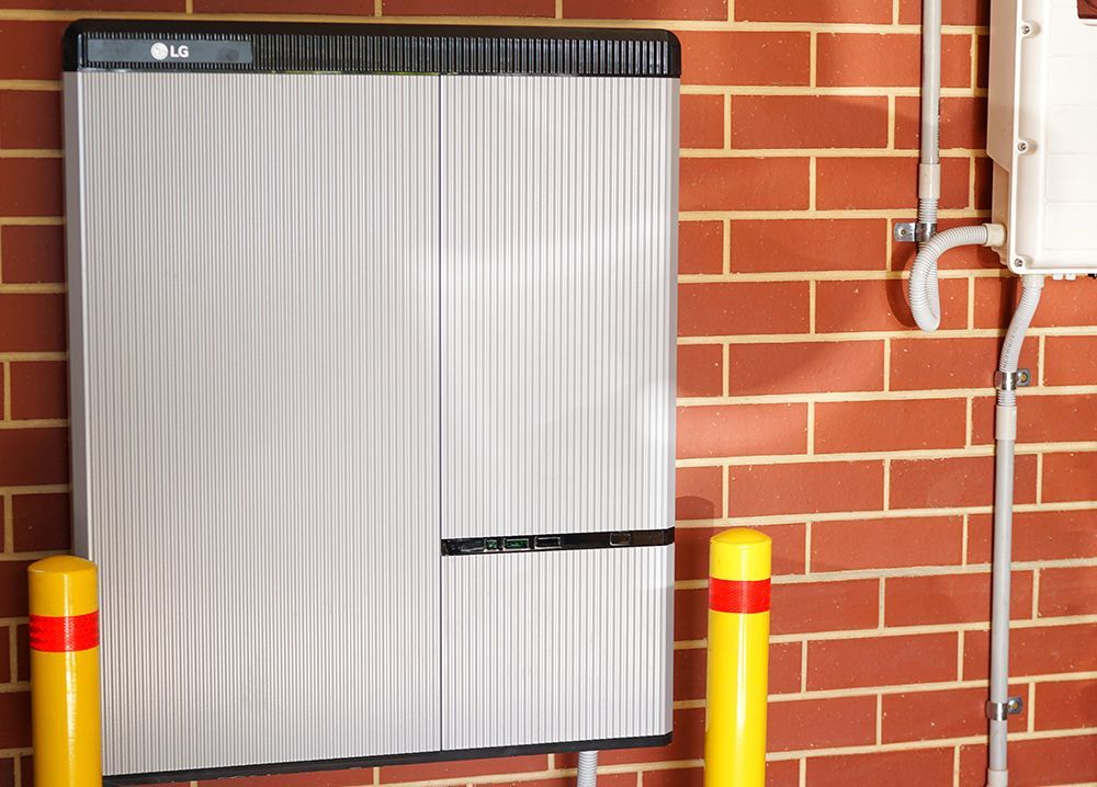 LG battery storage unit mounted on a brick wall, with yellow bollards and electrical conduit.