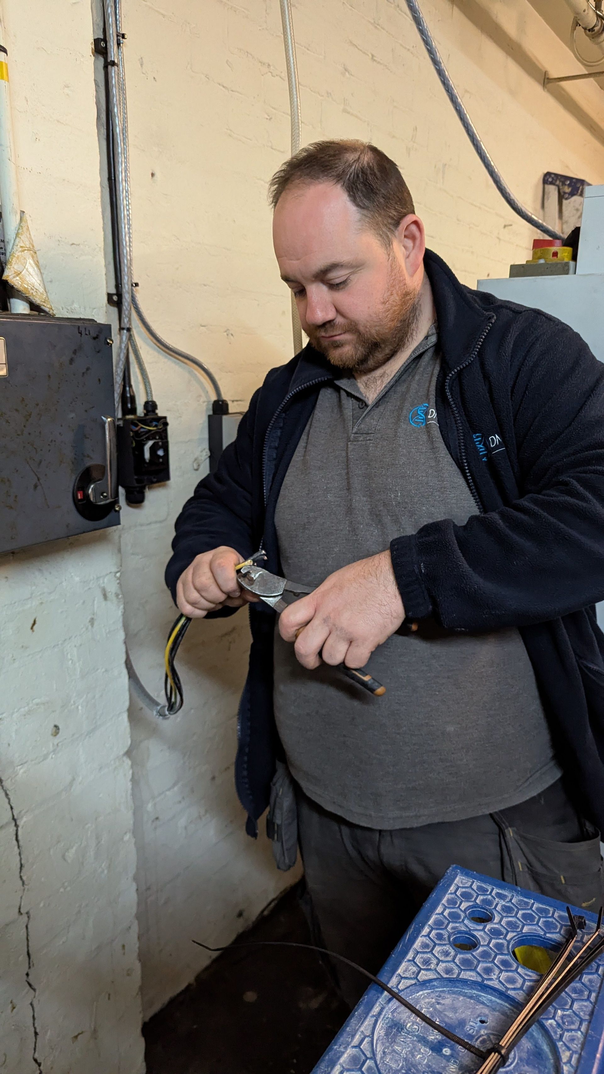 Man adjusting a pipe or tool in a utility room, with wall-mounted electrical boxes and cables nearby