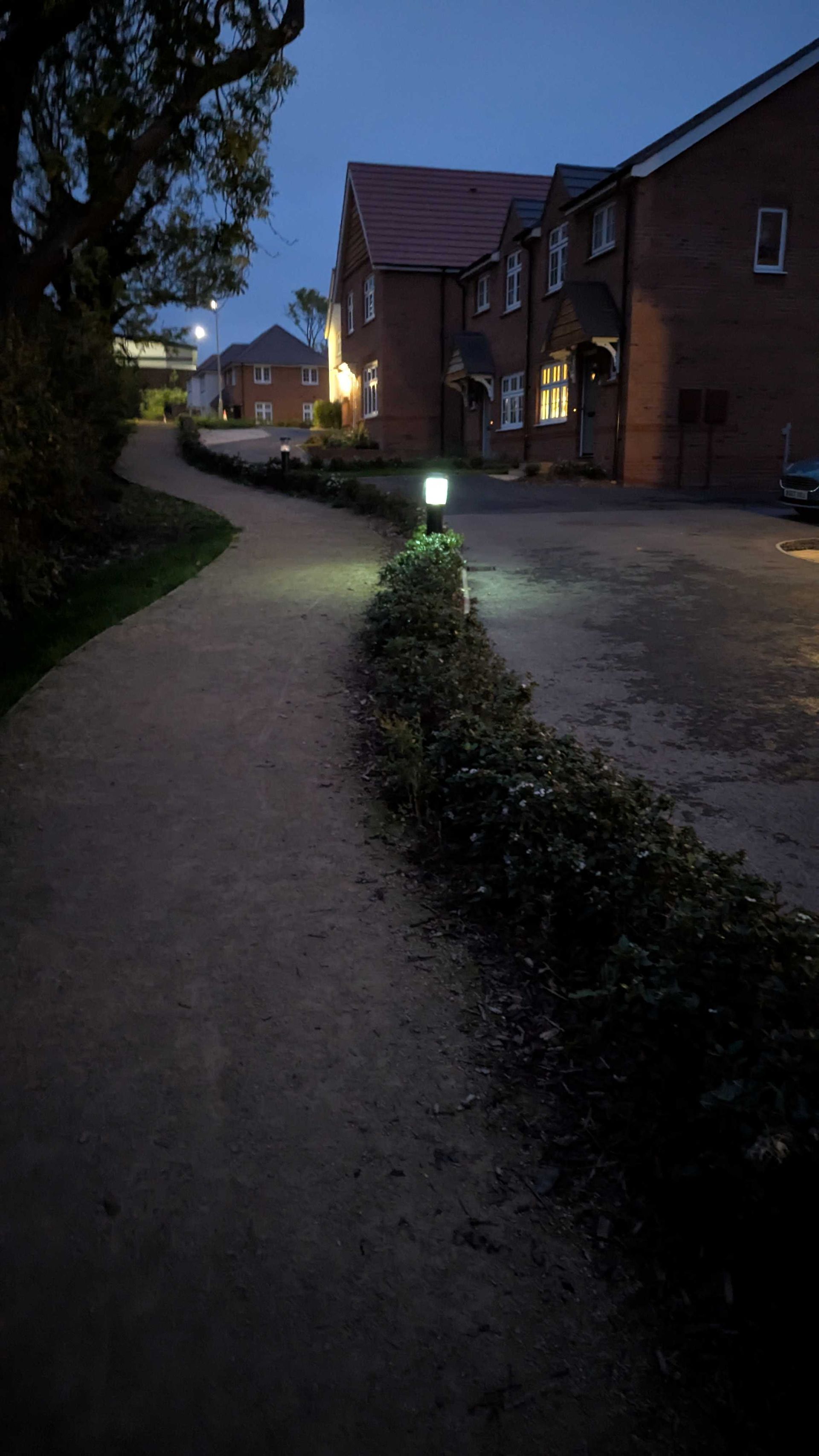 A gravel path winds past a hedge and houses at dusk, illuminated by a few lights.