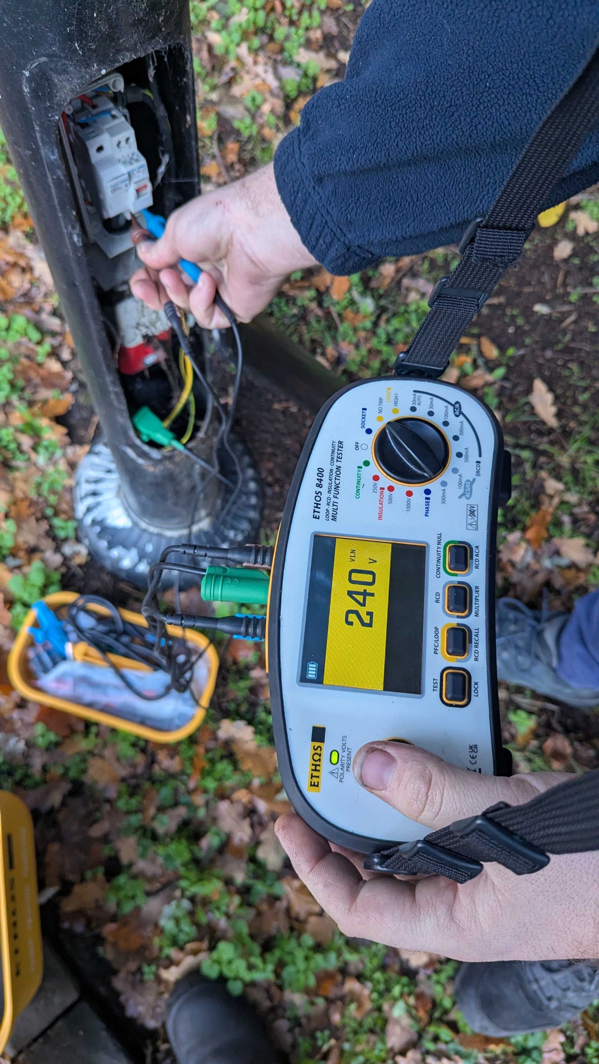 Person testing electrical wires in a utility pole with a multimeter; display reads 240.