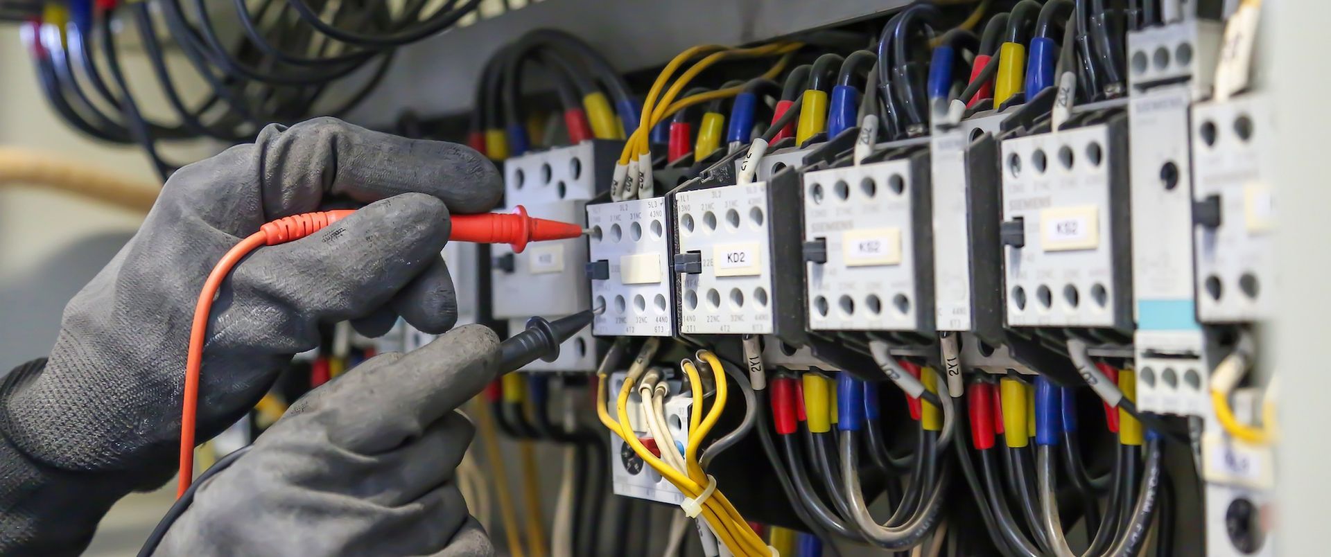 Electrician wearing gloves tests wires in a control panel with a multimeter.