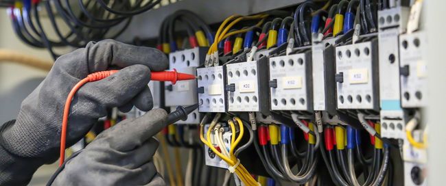 Electrician wearing gloves tests wires in a control panel with a multimeter.